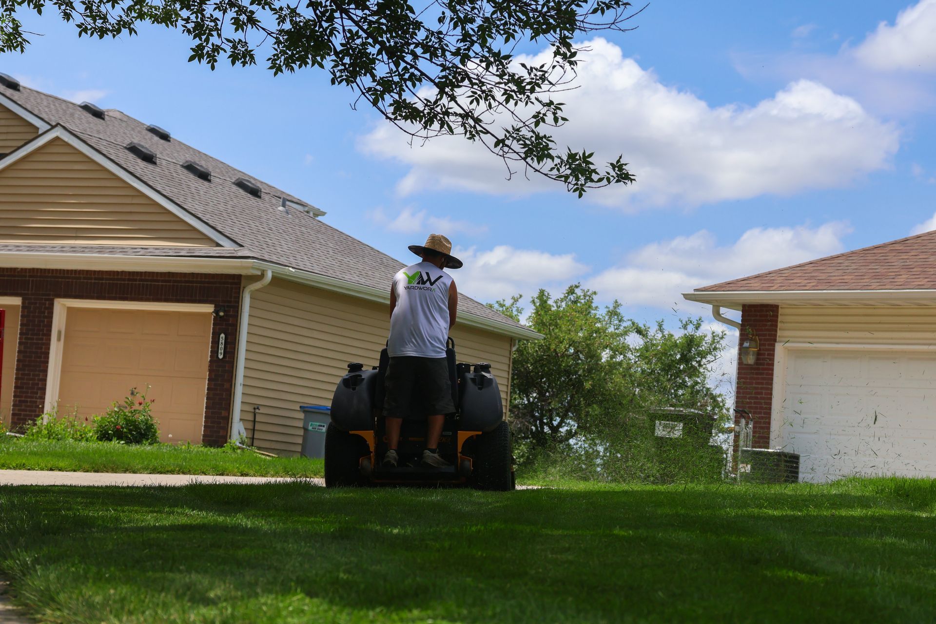 Person on lawnmower cutting grass in front of houses on a sunny day.