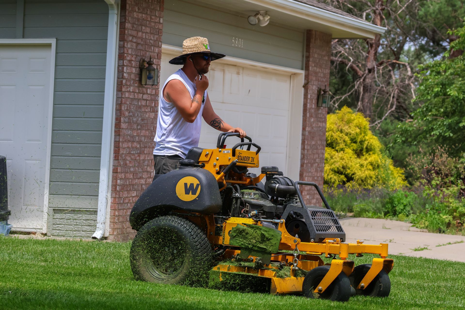 A person mowing a lawn with a yellow commercial riding mower in front of a house.