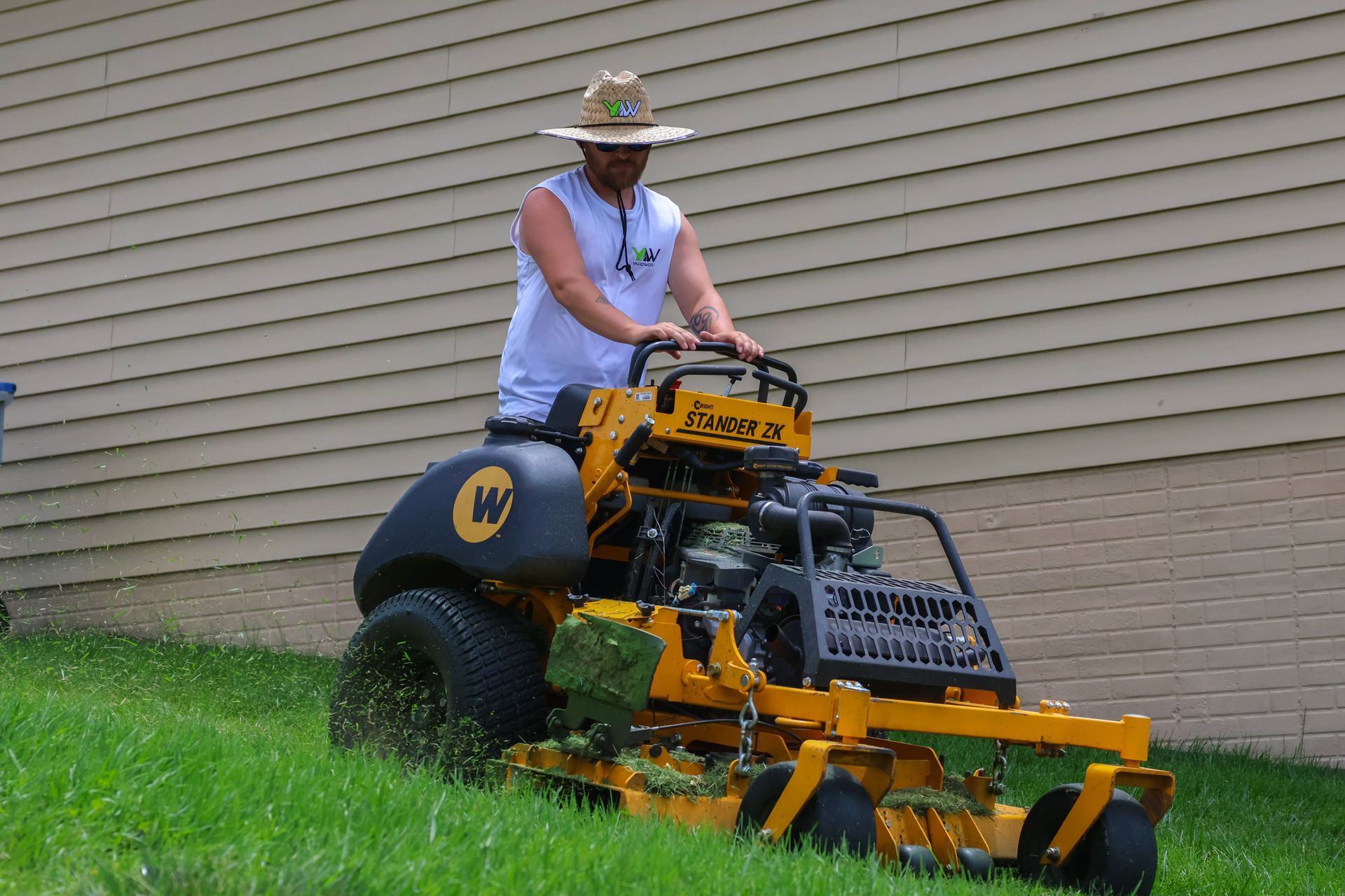Man on yellow lawnmower cutting grass on a sloped yard near a light brown house.