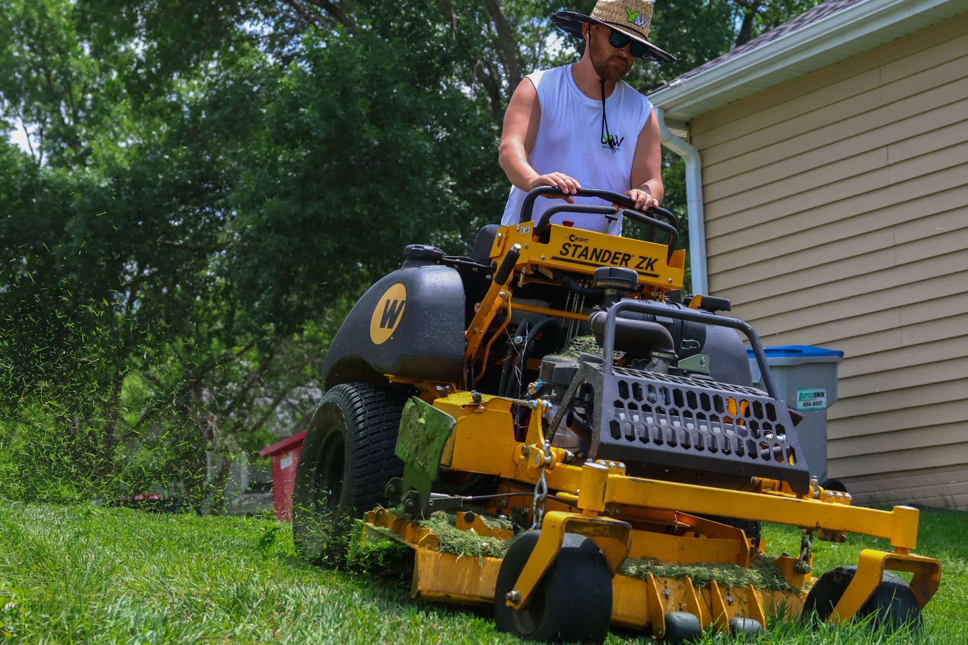 Man on a yellow zero-turn mower cutting grass near a house on a sunny day.