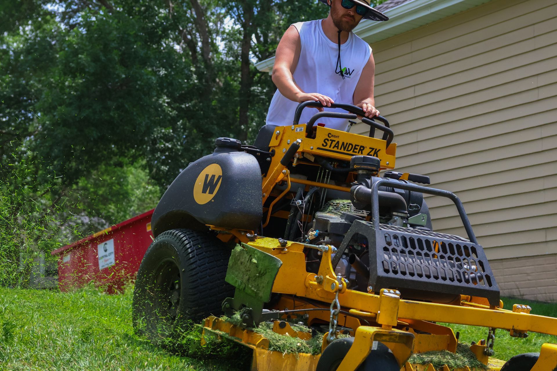A person mowing grass on a yellow zero-turn mower; green lawn and red debris container are visible.