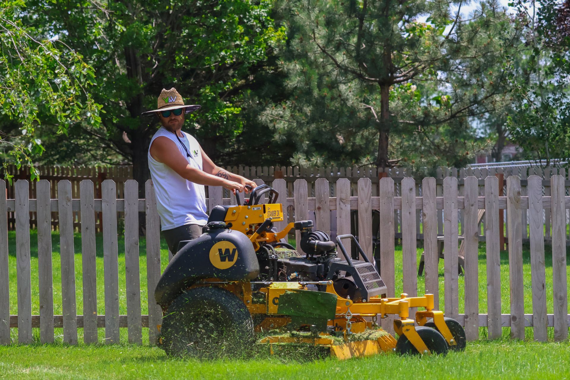 Man mowing a lawn with a yellow commercial mower near a white picket fence and trees.
