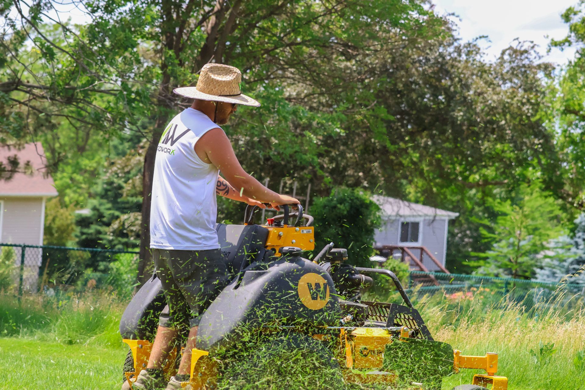 Man mowing grass on a yellow lawnmower, wearing a straw hat and white tank top.