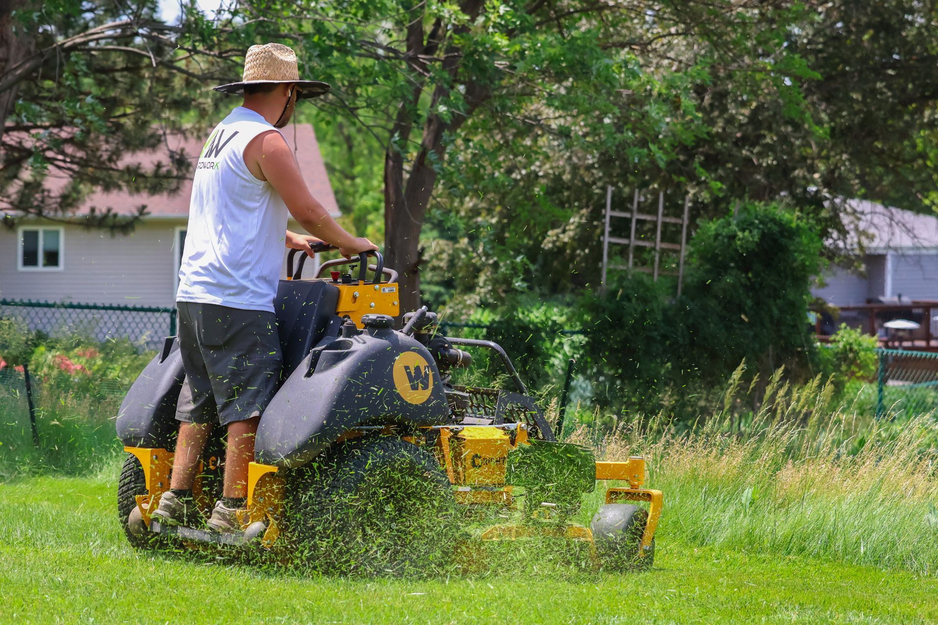 Person mowing a lawn on a yellow and black riding mower, grass flying up, sunny day.