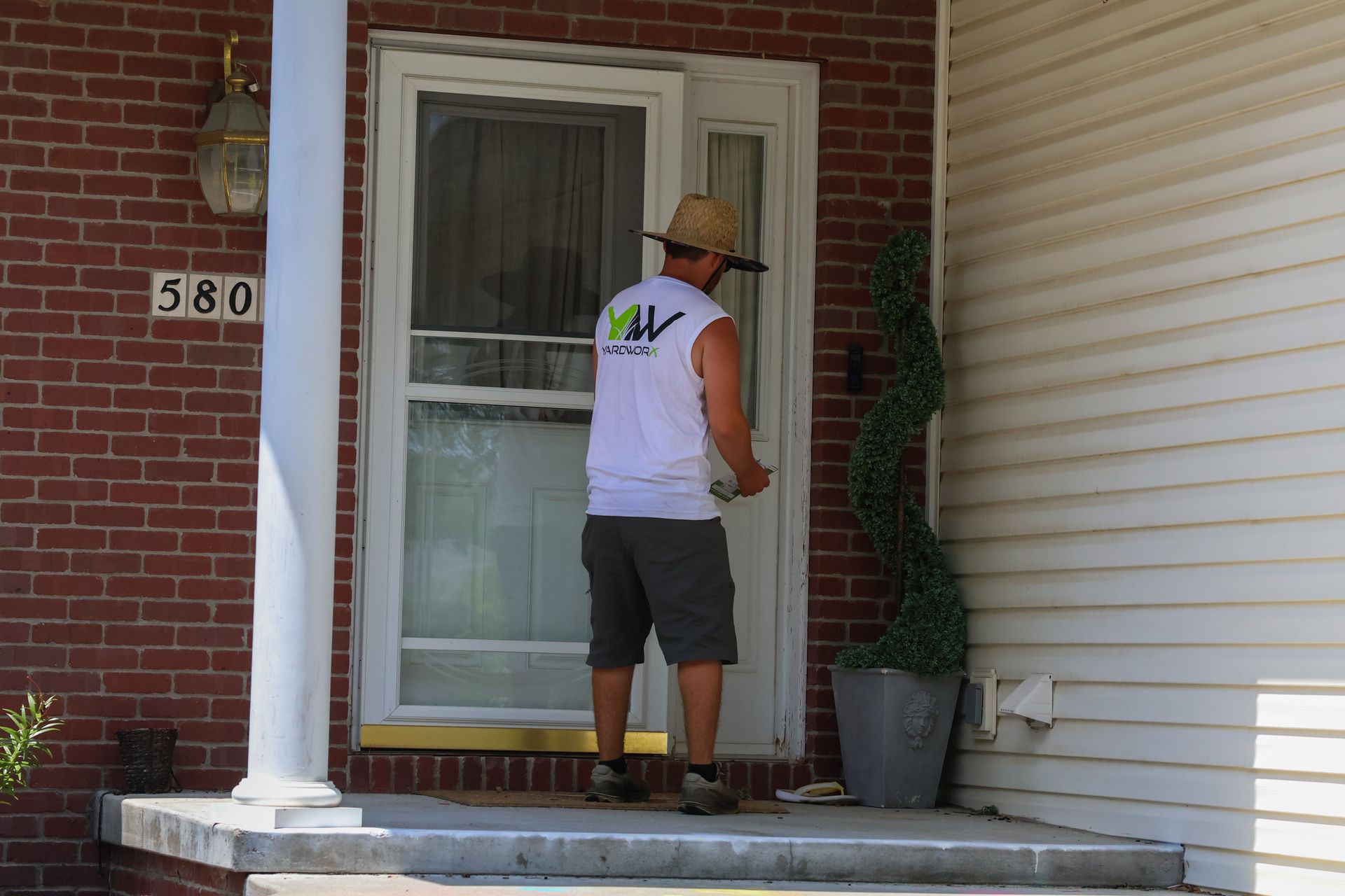 Person in work clothes at a door, holding something. House number 580 on brick. White door, column, and siding.