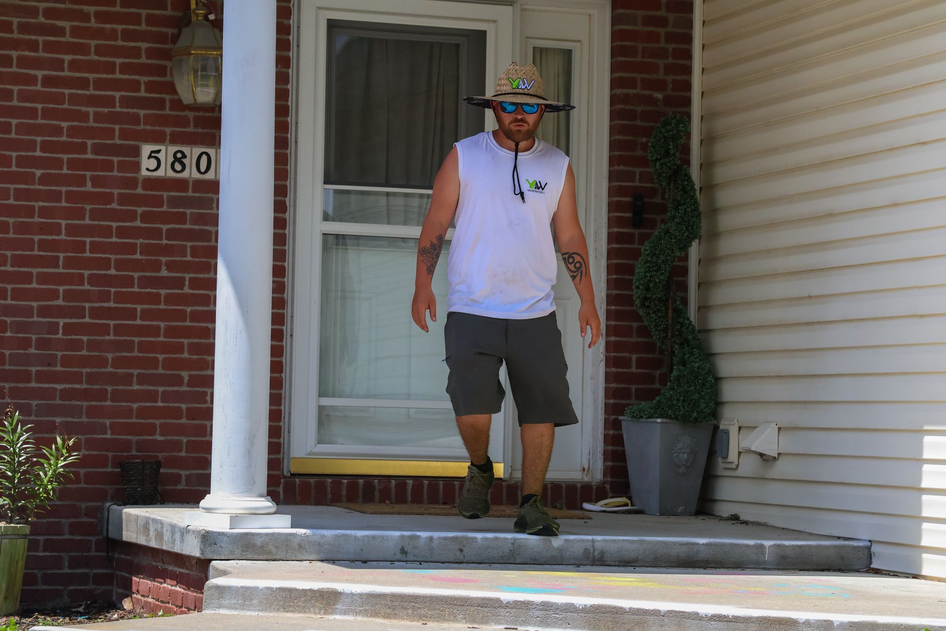 Man exiting a house with a straw hat and cargo shorts. The house is brick with a white porch.