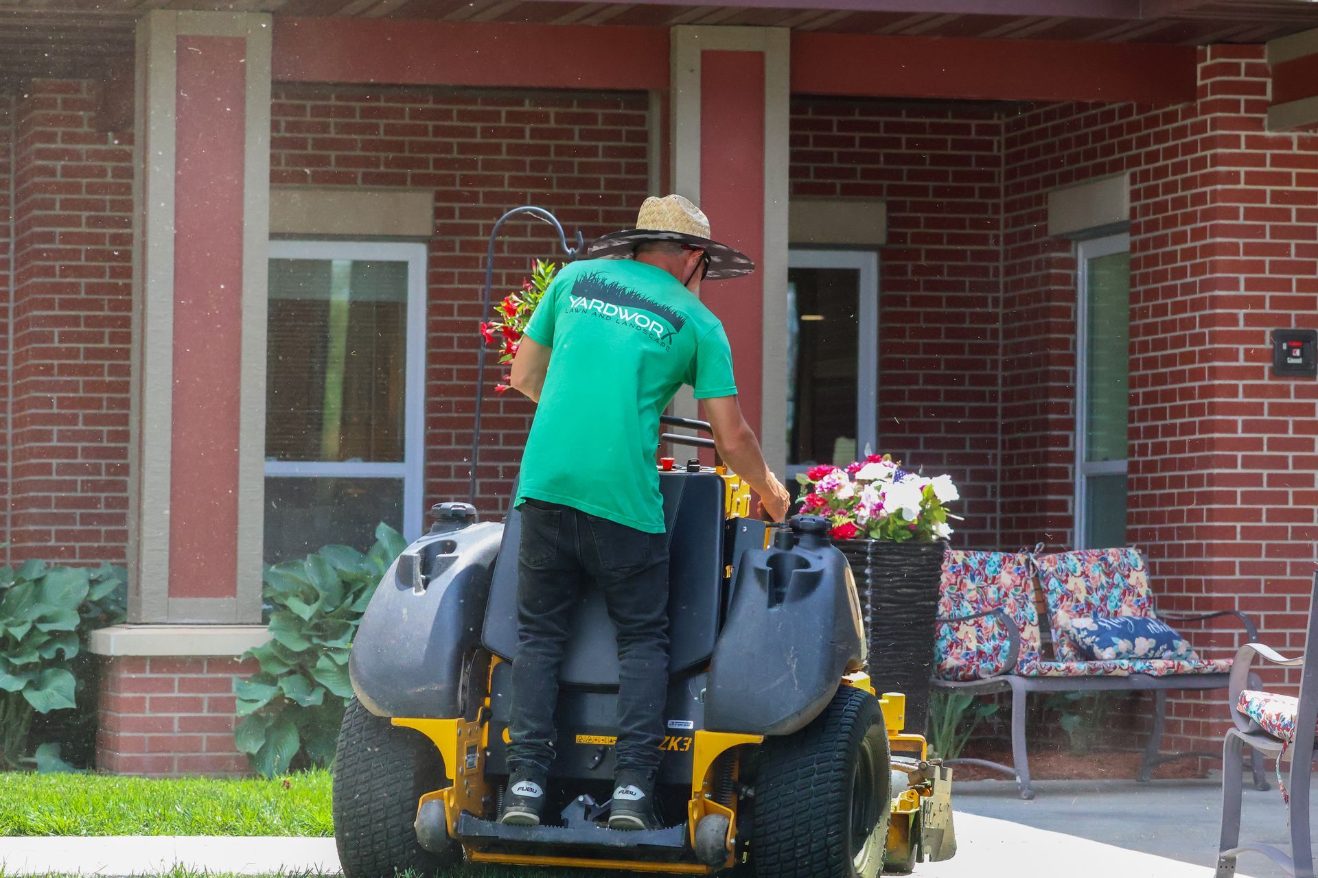 Man on a riding mower with hanging flower baskets; in front of a brick building.