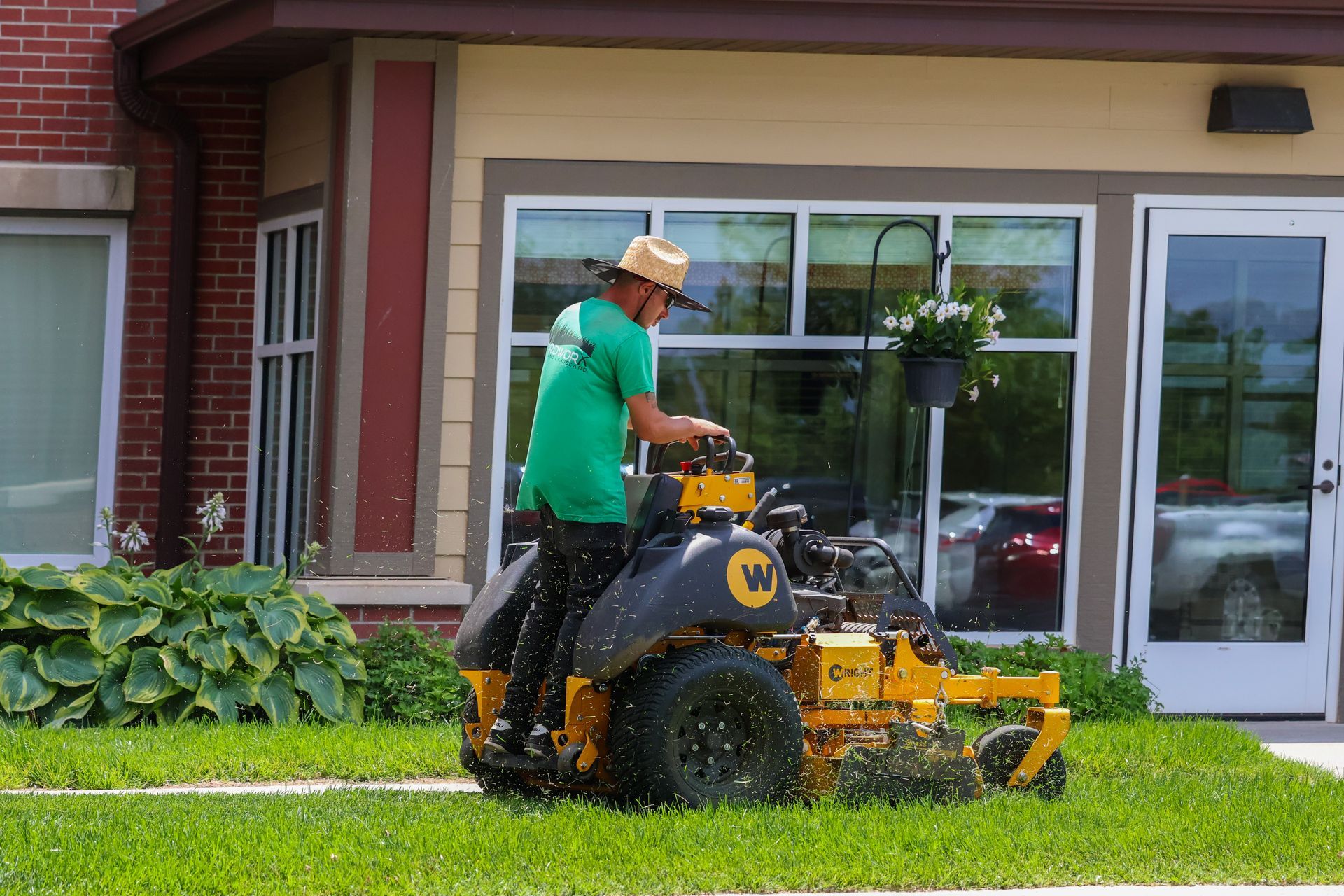 Person mowing grass with a yellow lawnmower near a building with windows. Green shirt, straw hat.