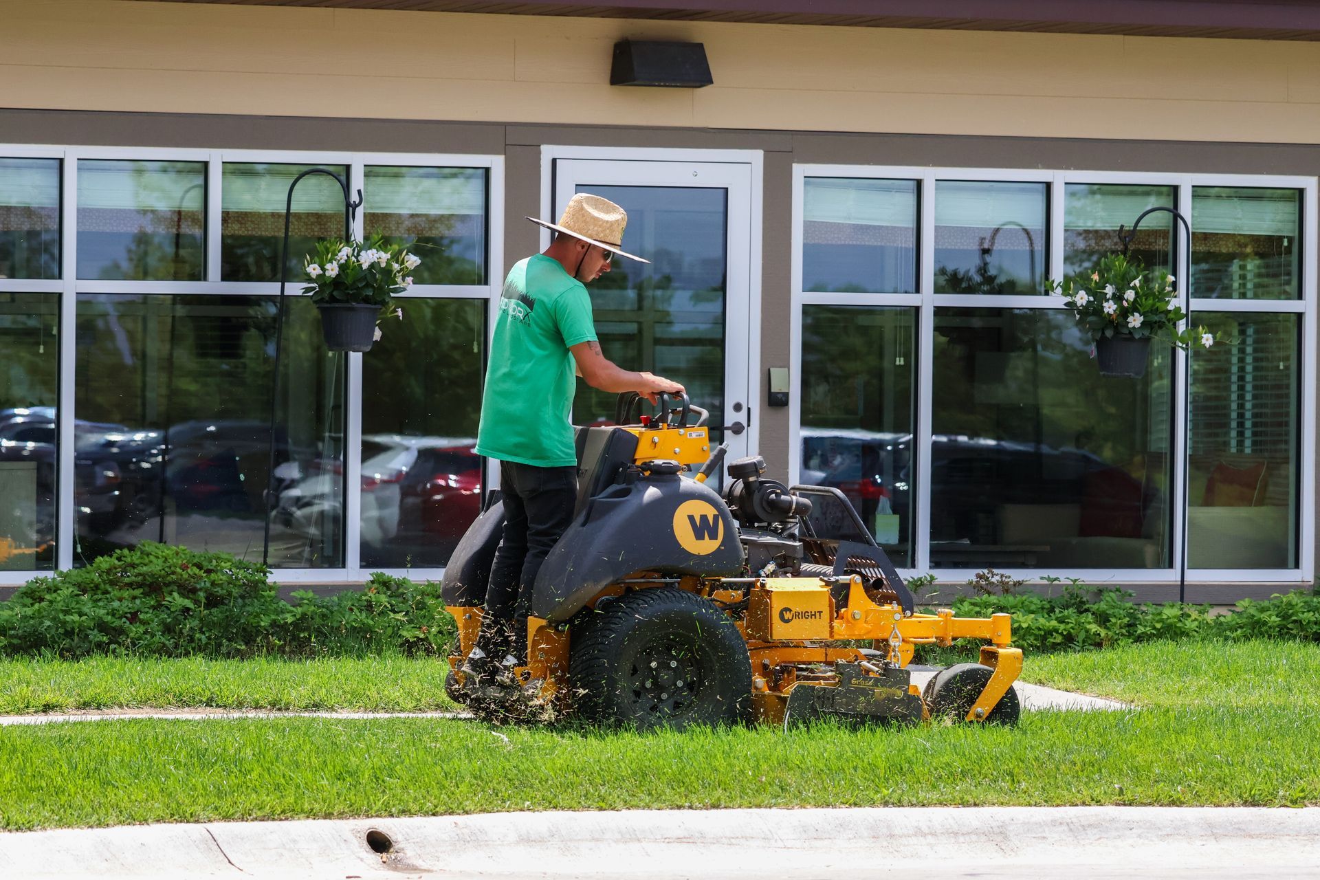 A person mowing grass with a yellow zero-turn mower in front of a building with large windows.