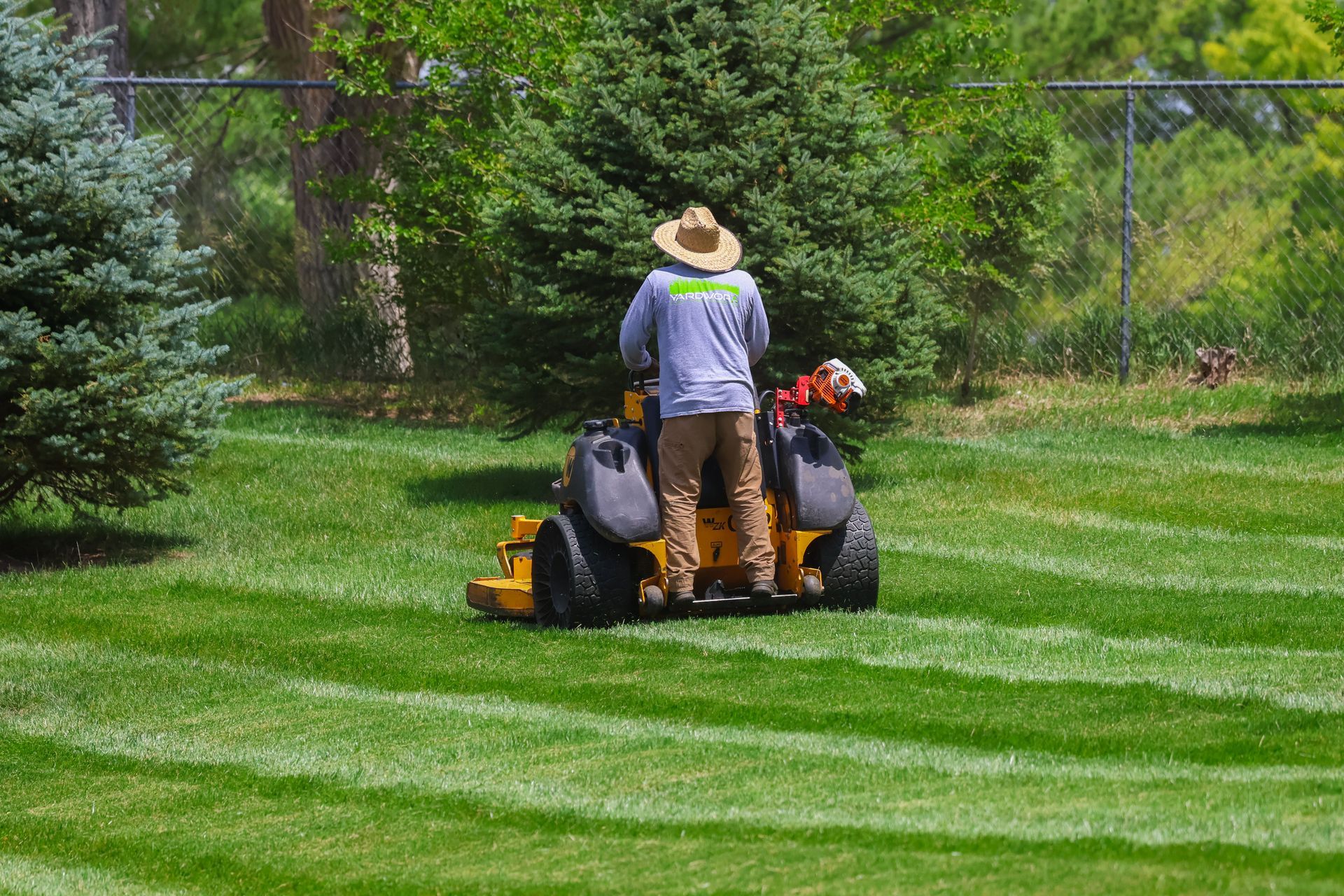 Person in straw hat mowing a green lawn with a yellow riding mower on a sunny day.