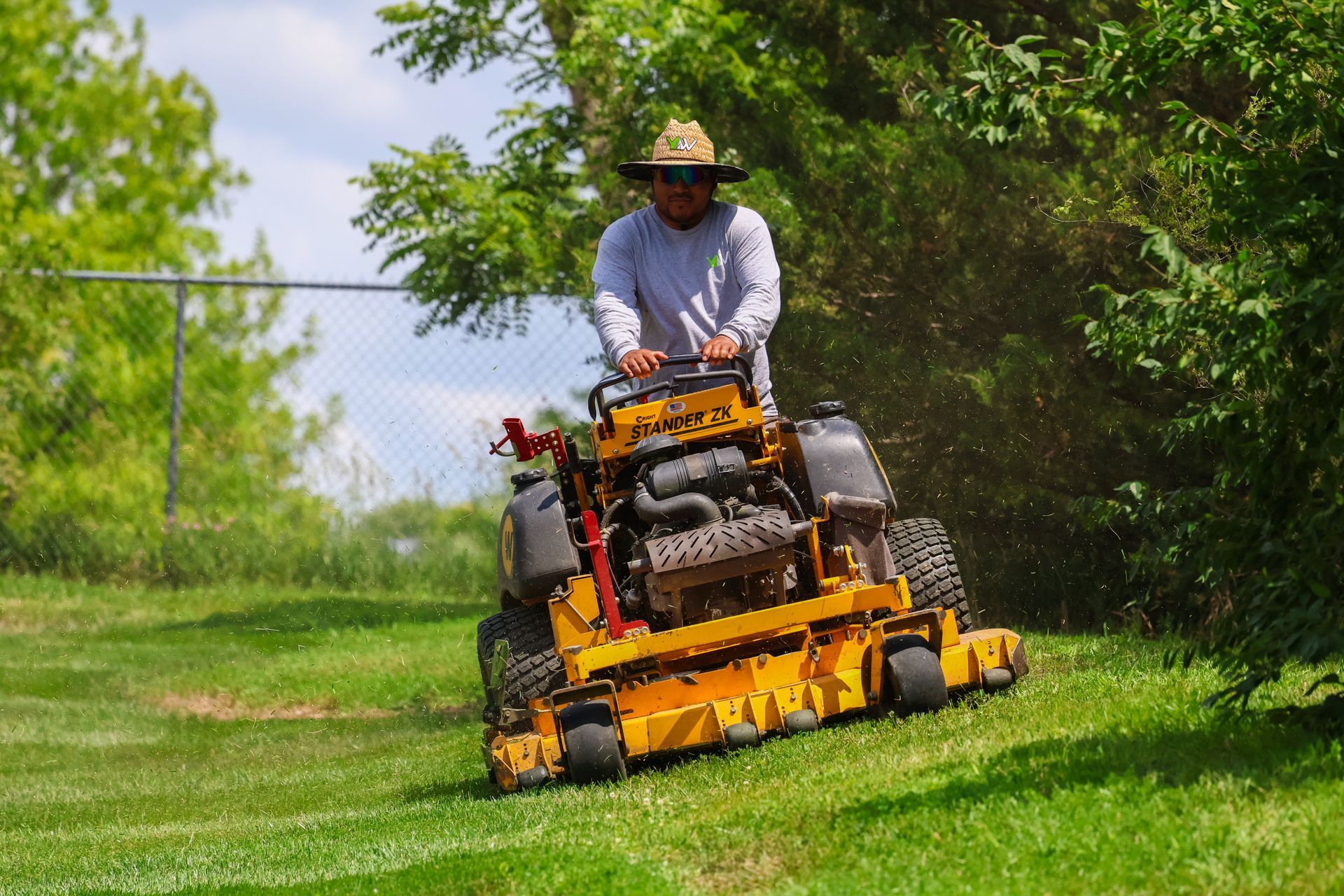 Person mowing grass on a yellow zero-turn mower on a sloped, grassy area near trees.