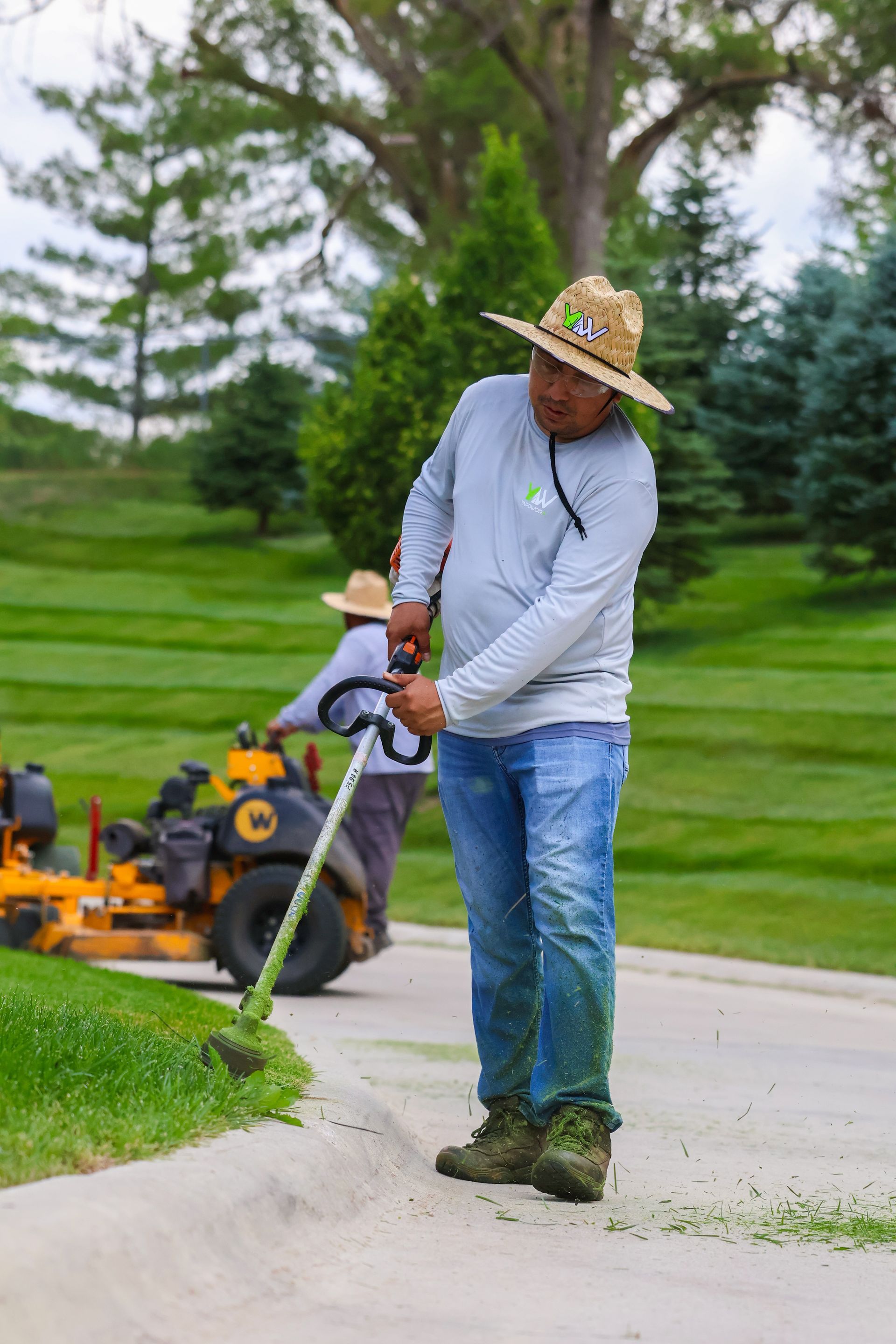 Man wearing straw hat using string trimmer along a curb, other worker and mower in background.