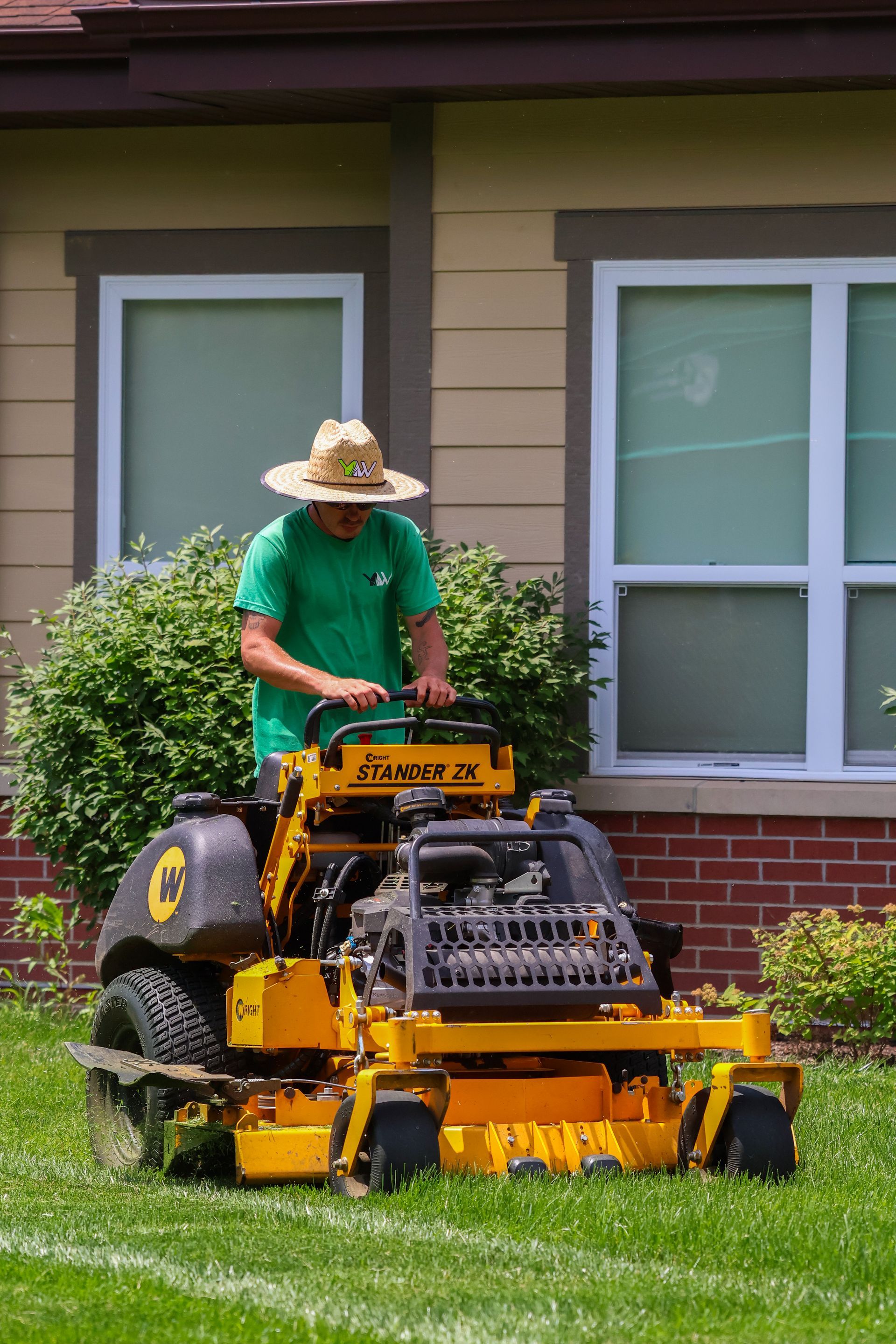 Person mowing a lawn with a yellow zero-turn mower in front of a house.