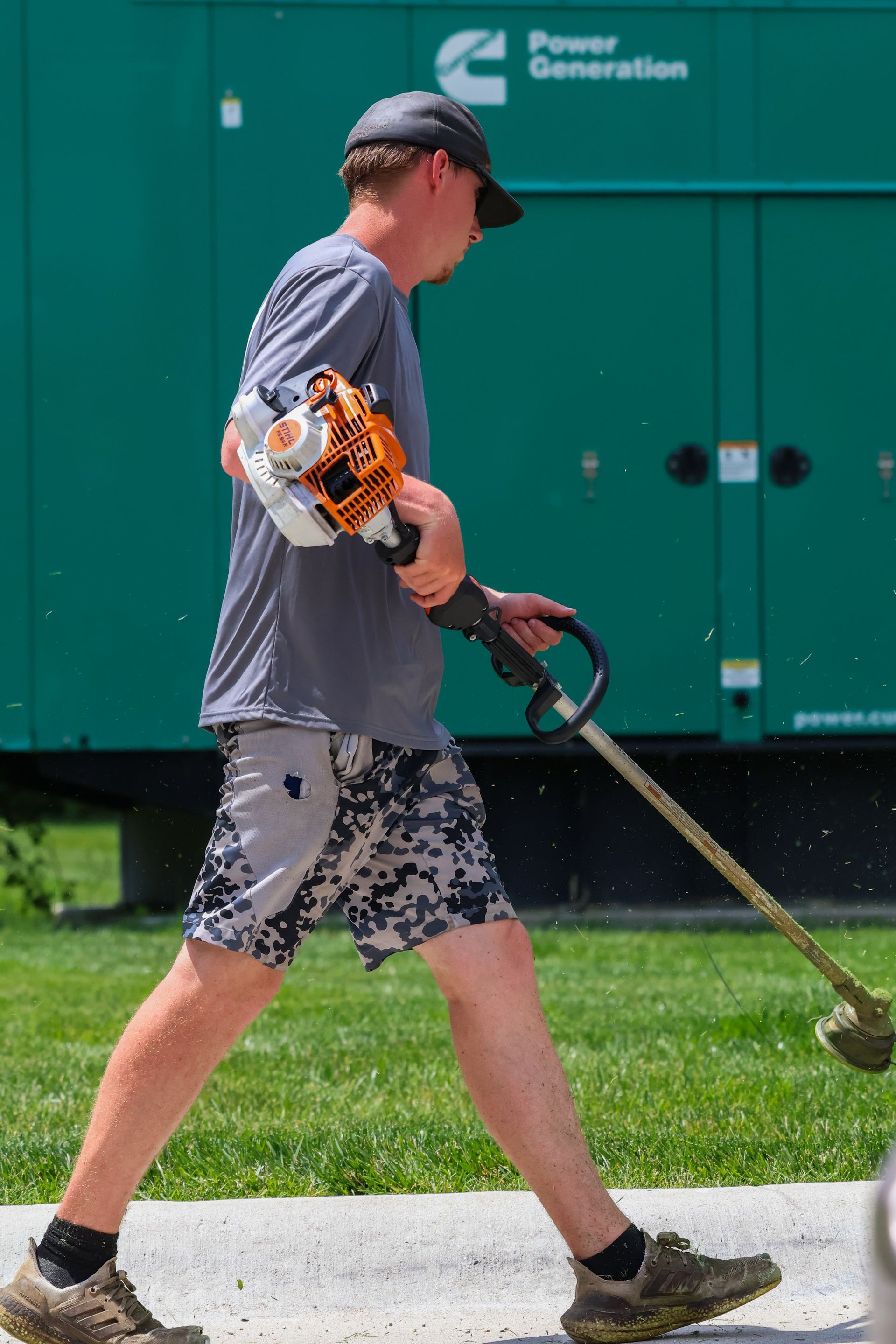 Man using a weed whacker outdoors near a green generator. He wears shorts, a shirt, cap, and work boots.