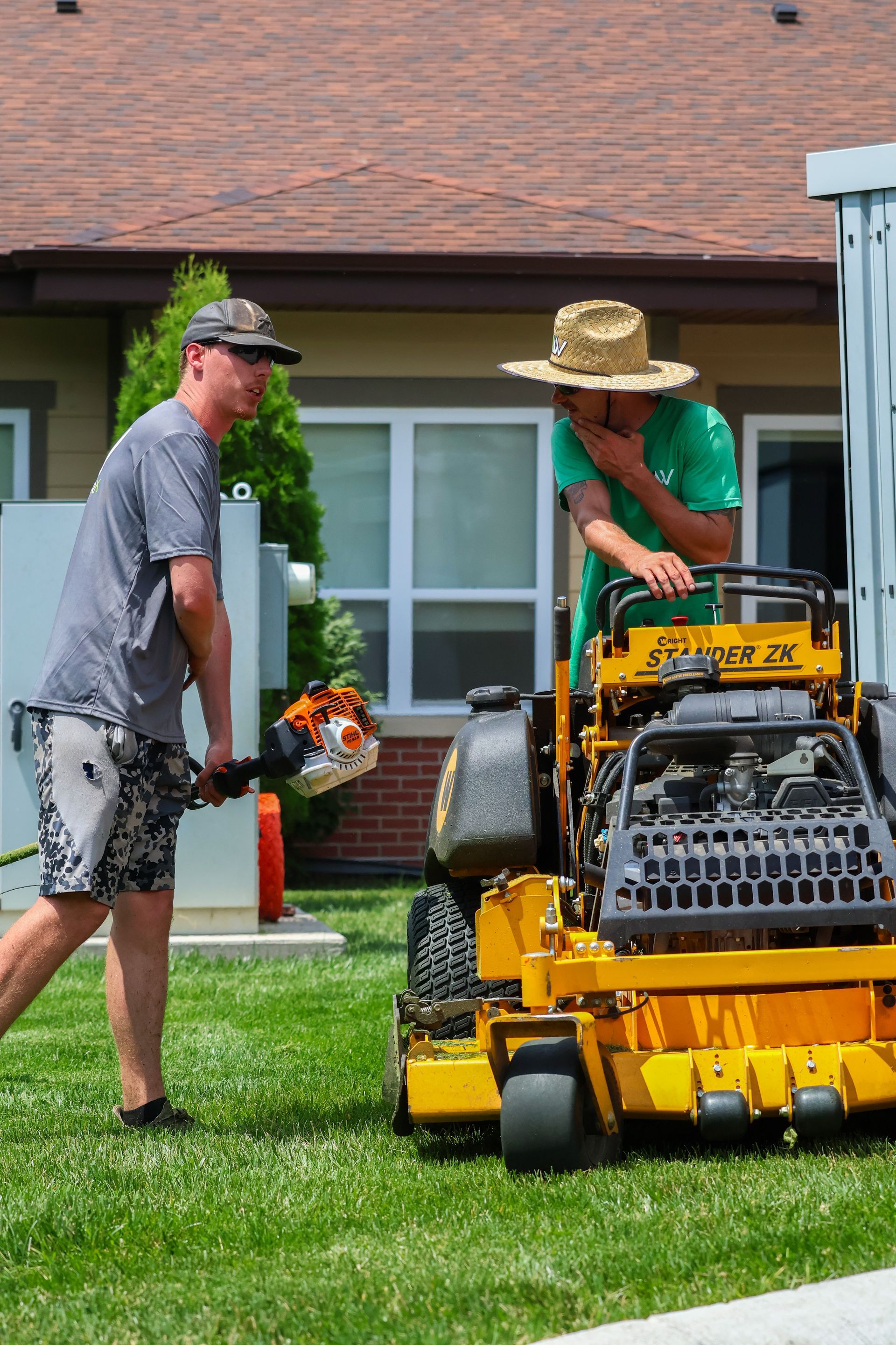 Two people mowing lawn: one with a trimmer, the other on a yellow riding mower. Sunny outdoors with a building backdrop.