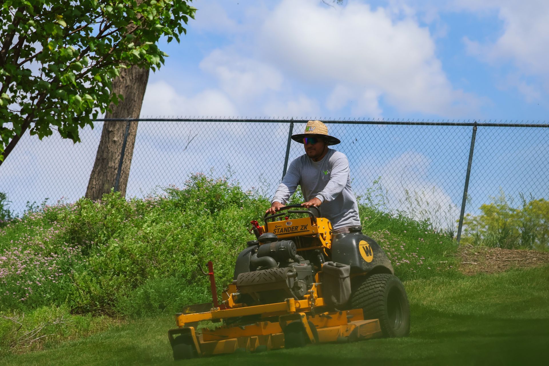 Person wearing straw hat operating a yellow lawn mower on a grassy field; blue sky background.