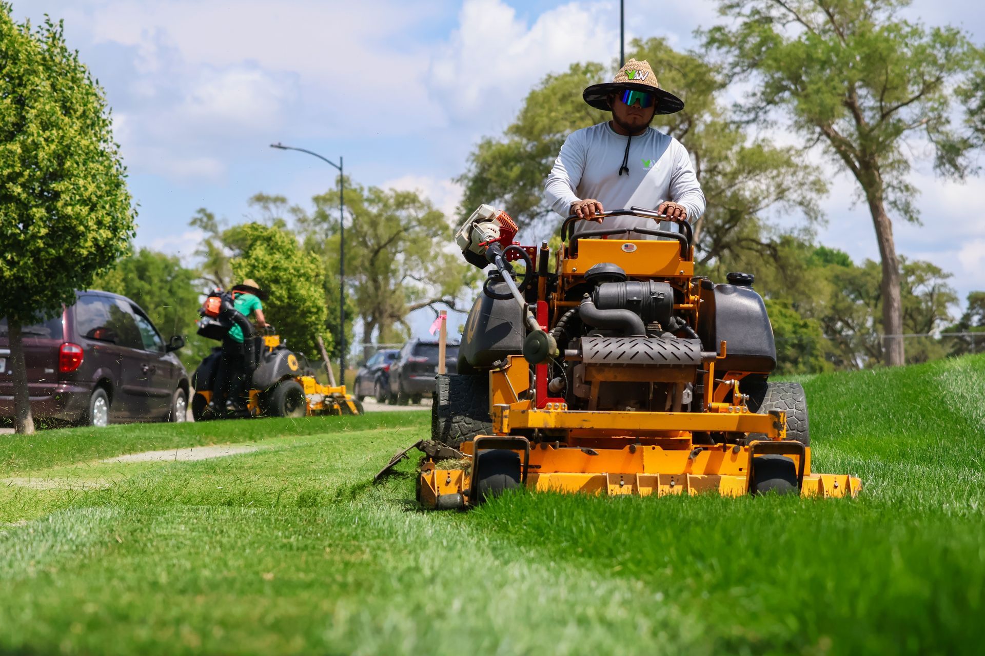 Man operating a yellow zero-turn mower, cutting grass in a sunny park with other mowers nearby.