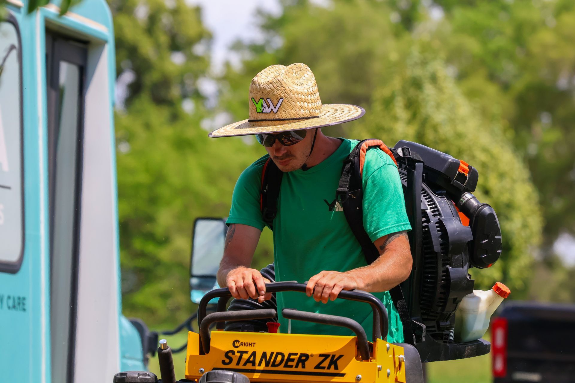 Man operating a lawnmower, wearing a straw hat and backpack blower. The machine is yellow, and the setting is outdoors.