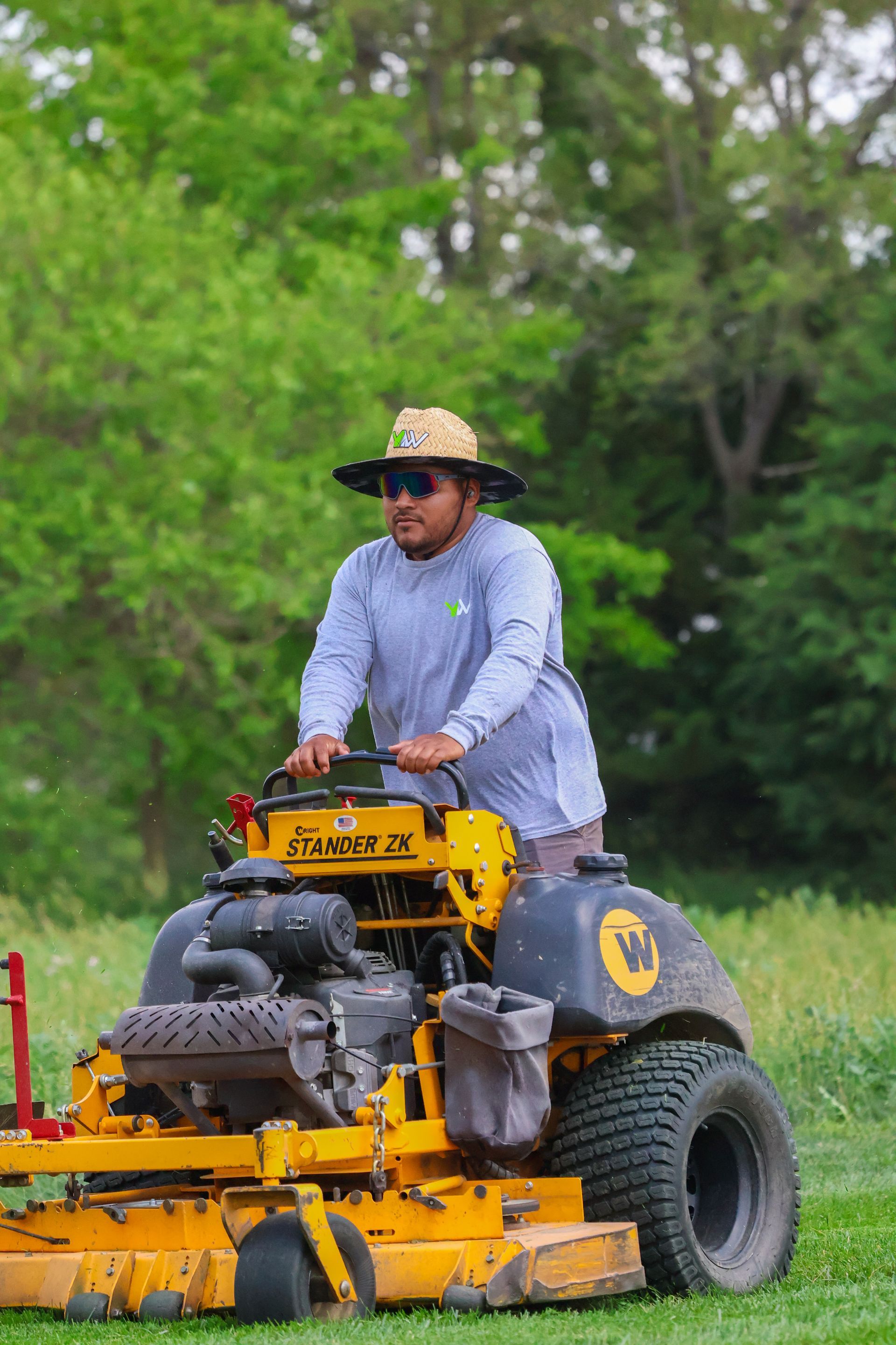 Person operating a yellow zero-turn mower in a grassy field, wearing a hat and long sleeves.