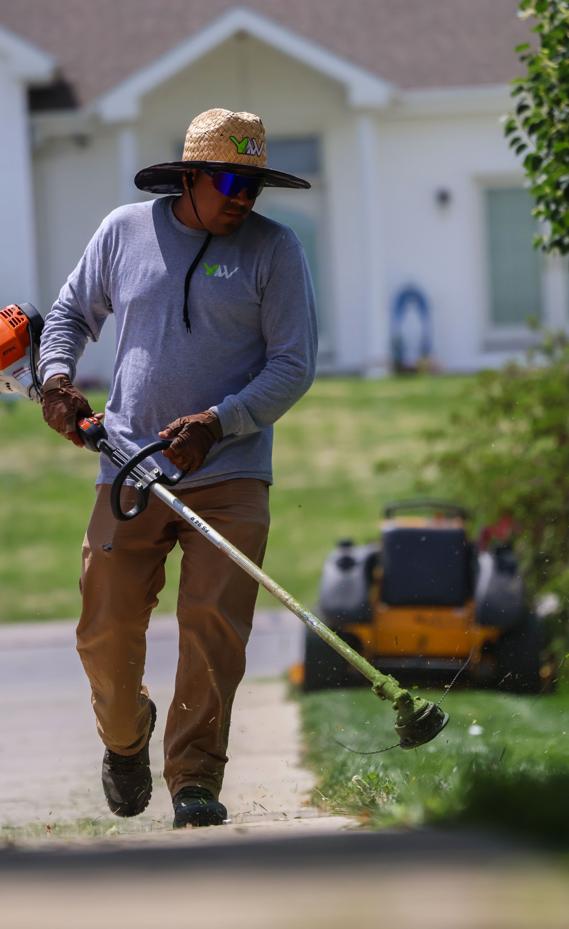 Landscaper using a weed trimmer to edge a sidewalk next to a lawn. He wears a hat, long sleeve shirt, and pants.