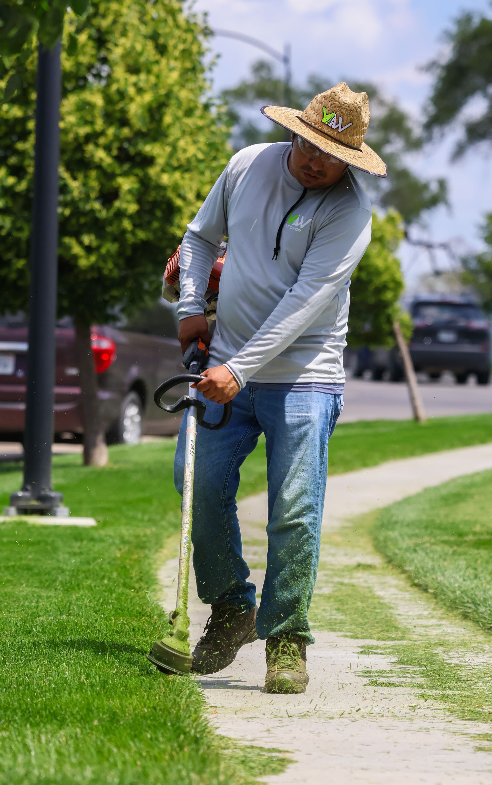 Person using a weed wacker to trim grass along a sidewalk. Wearing a hat, long-sleeved shirt, and jeans outdoors.