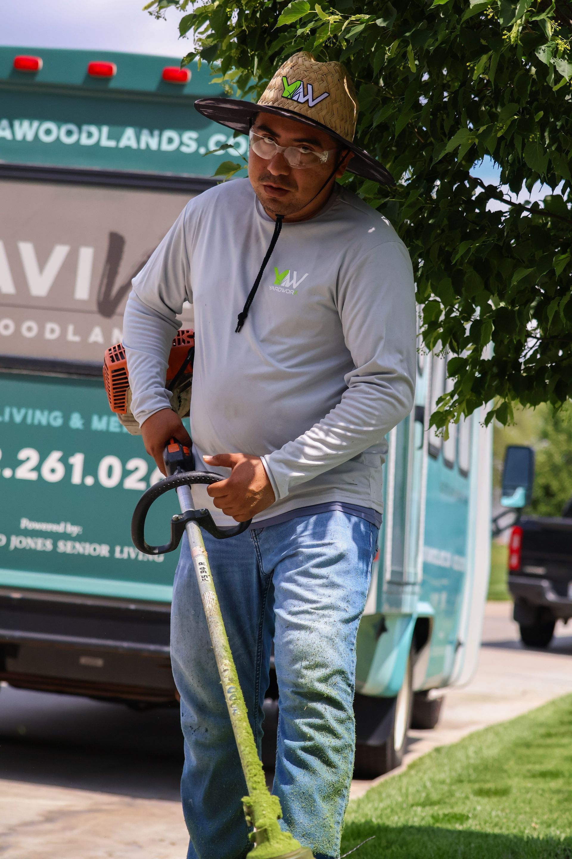 Man wearing straw hat using a string trimmer in front of a green vehicle.