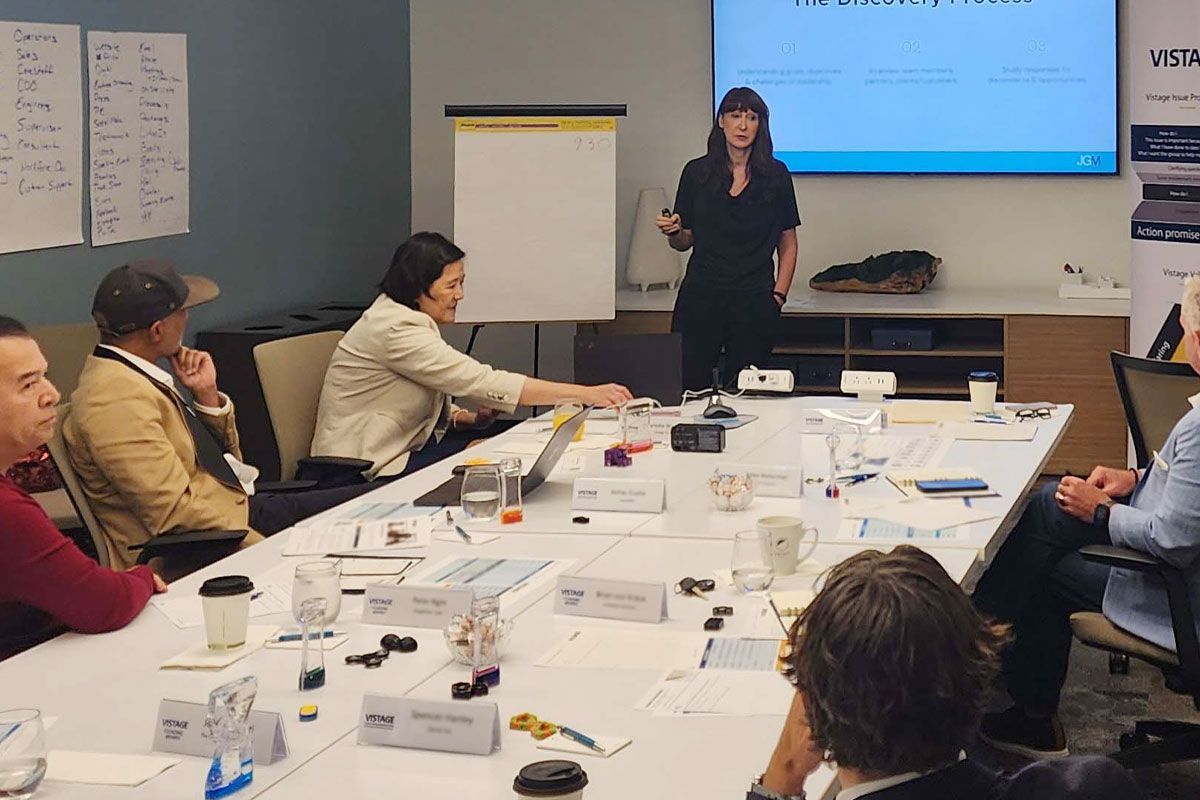Meeting room with people seated around a table while a presenter speaks at a screen.