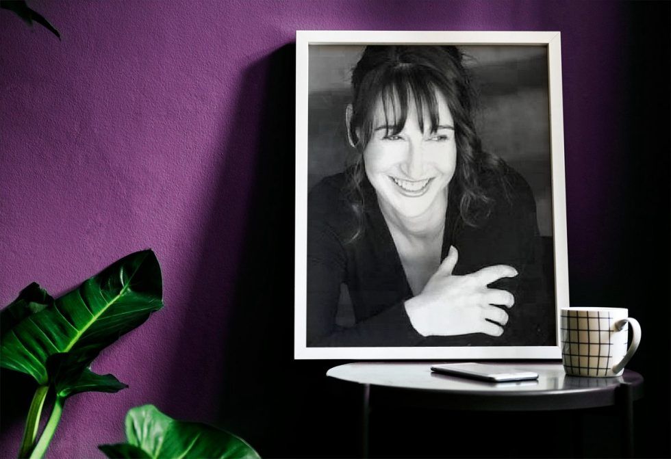 Black-and-white portrait on a small table beside a green plant against a purple wall, with a smiling woman