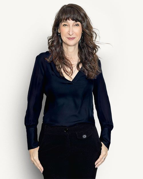 Woman in a dark blouse and black skirt standing against a light background.