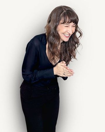 Smiling woman in a black outfit leaning forward with clasped hands against a beige background