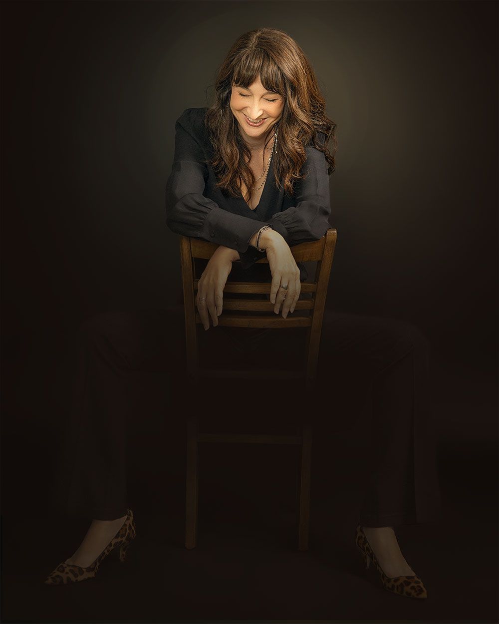 Woman leaning over a wooden chair in a dark studio, wearing black clothes and heels