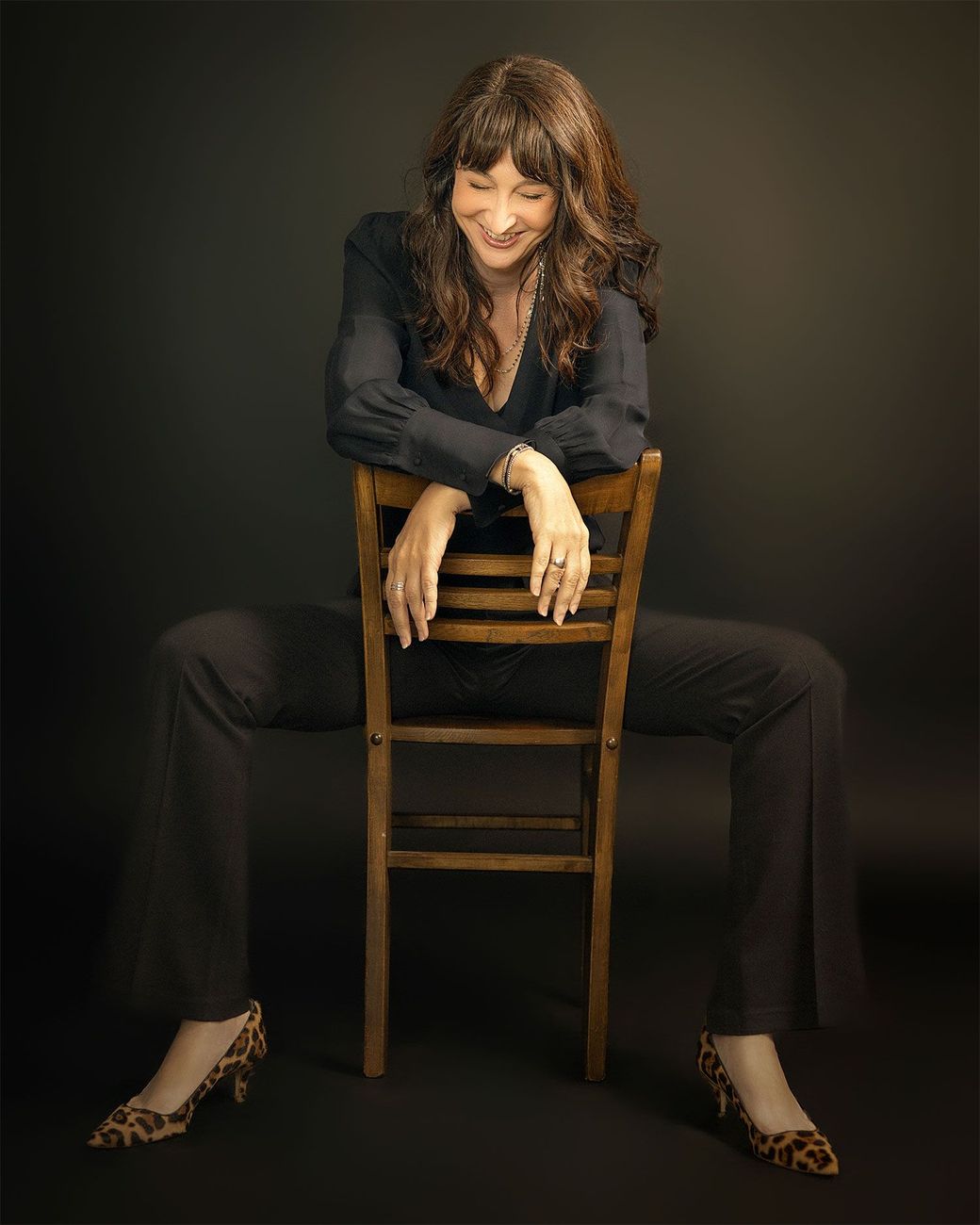 Woman leaning over a wooden chair in a dark studio, wearing black clothes and heels