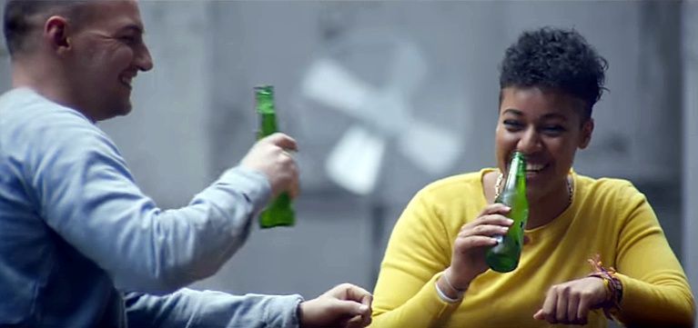 Two people smiling and holding green glass bottles in a casual indoor setting