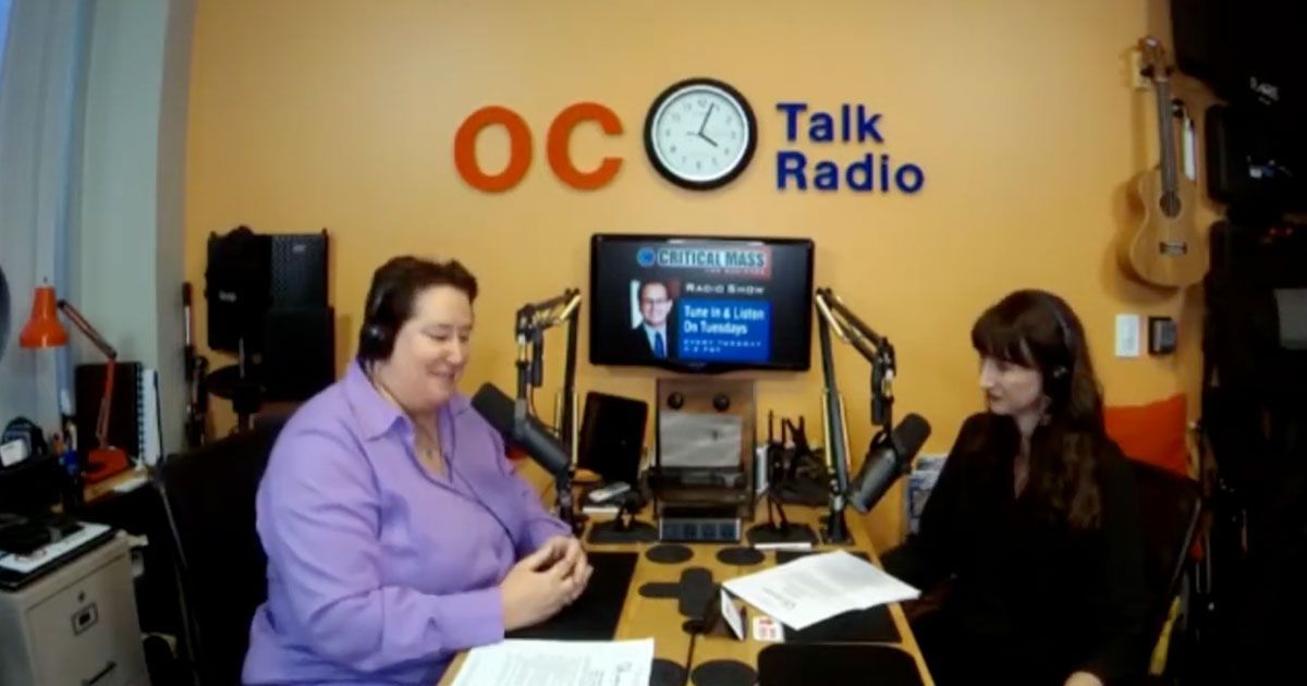 Two people recording a radio show in an OC Talk Radio studio, with microphones and a monitor behind them.