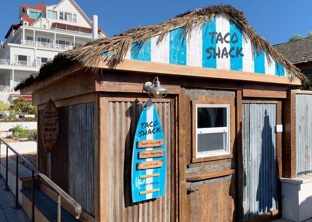 Small wooden taco shack with blue signs and a thatched roof on a boardwalk by the water