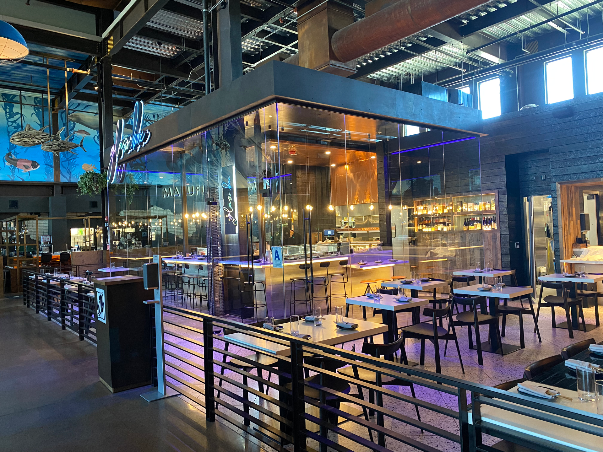 Modern indoor restaurant with glass walls, wooden tables, warm lighting, and an open kitchen/bar area