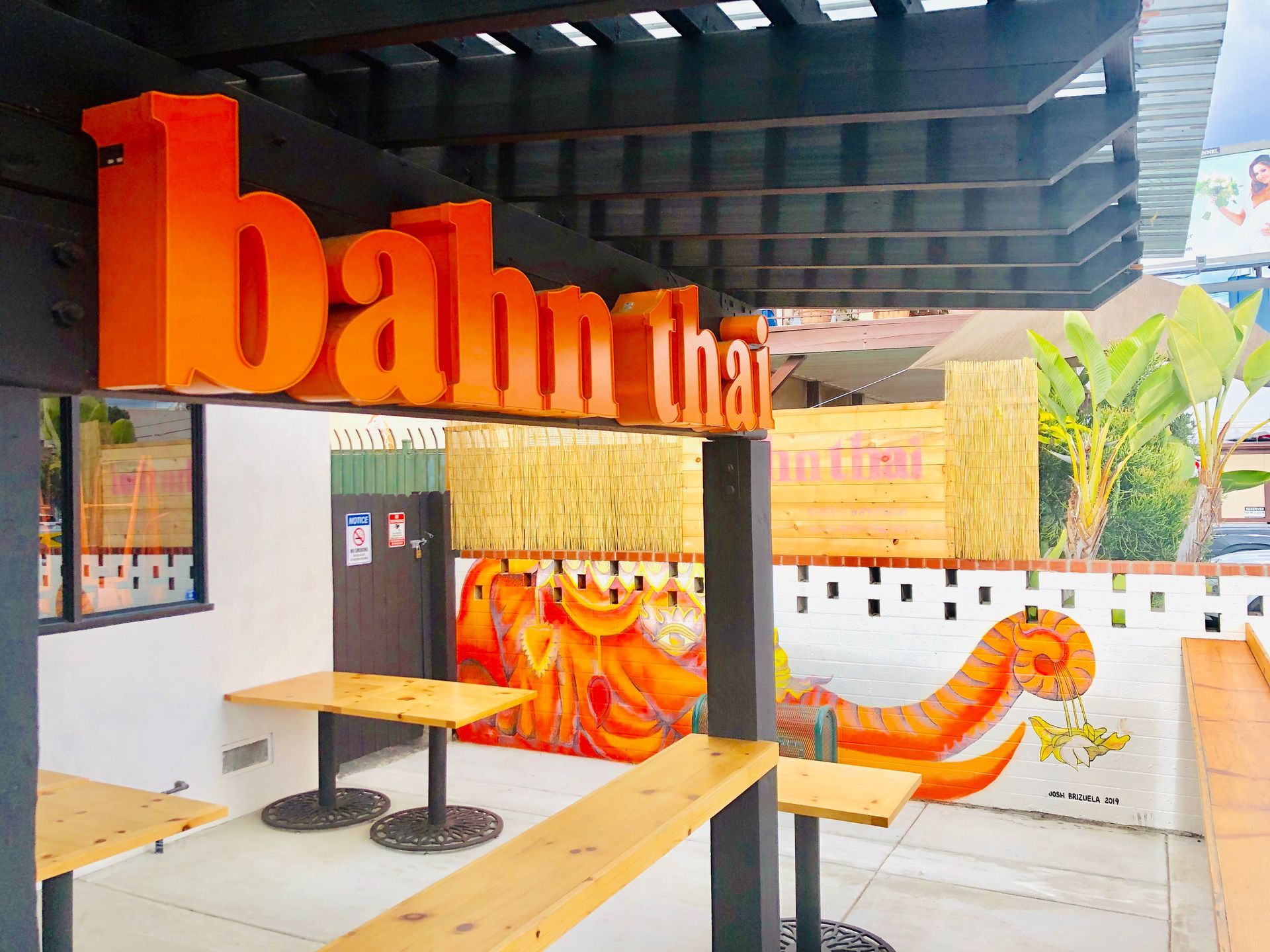 Orange “bahan thai” sign above a small outdoor seating area with wooden tables and a patterned wall mural