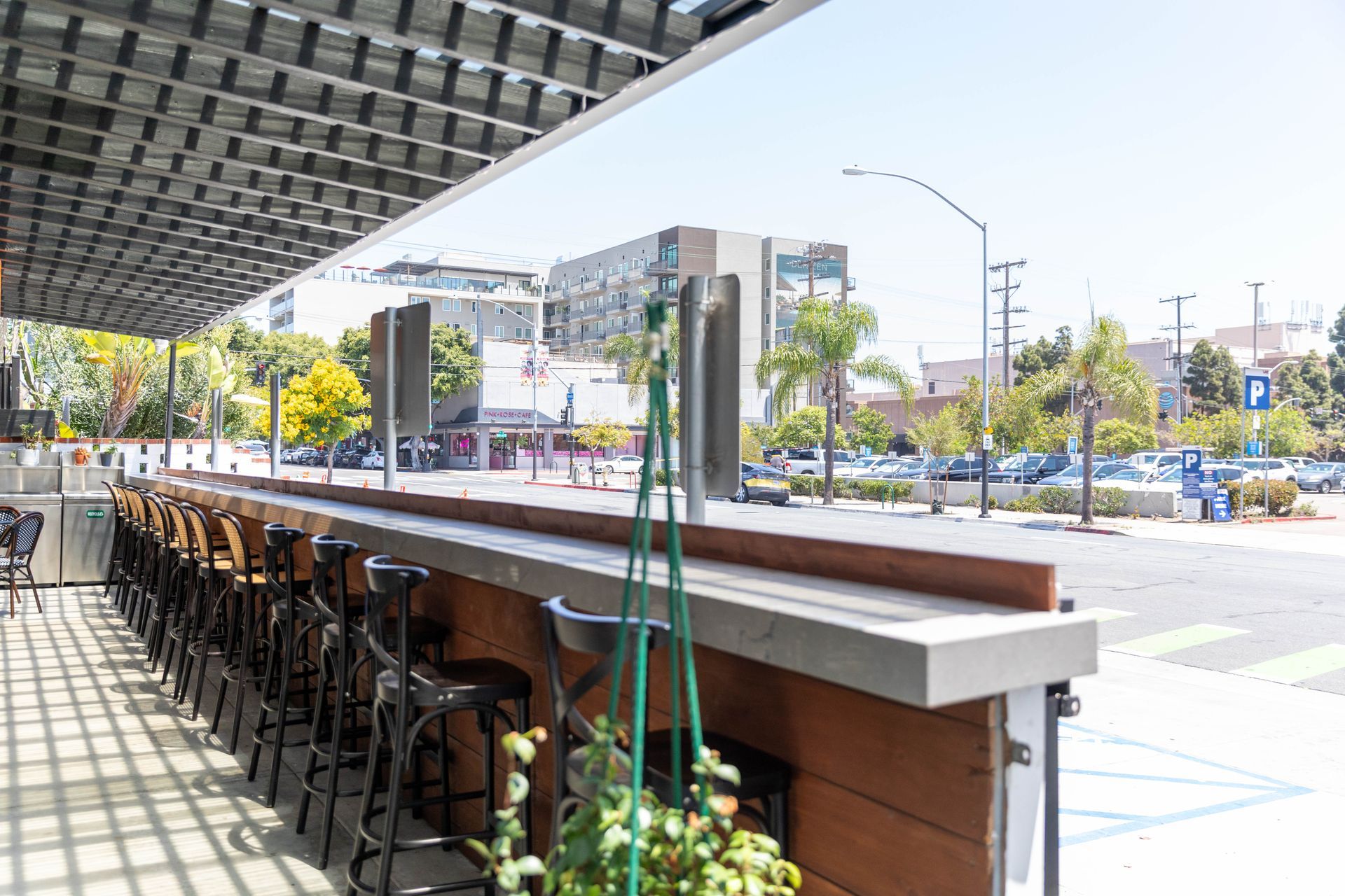 Outdoor café seating under an awning beside a sunny street with apartment buildings and trees.
