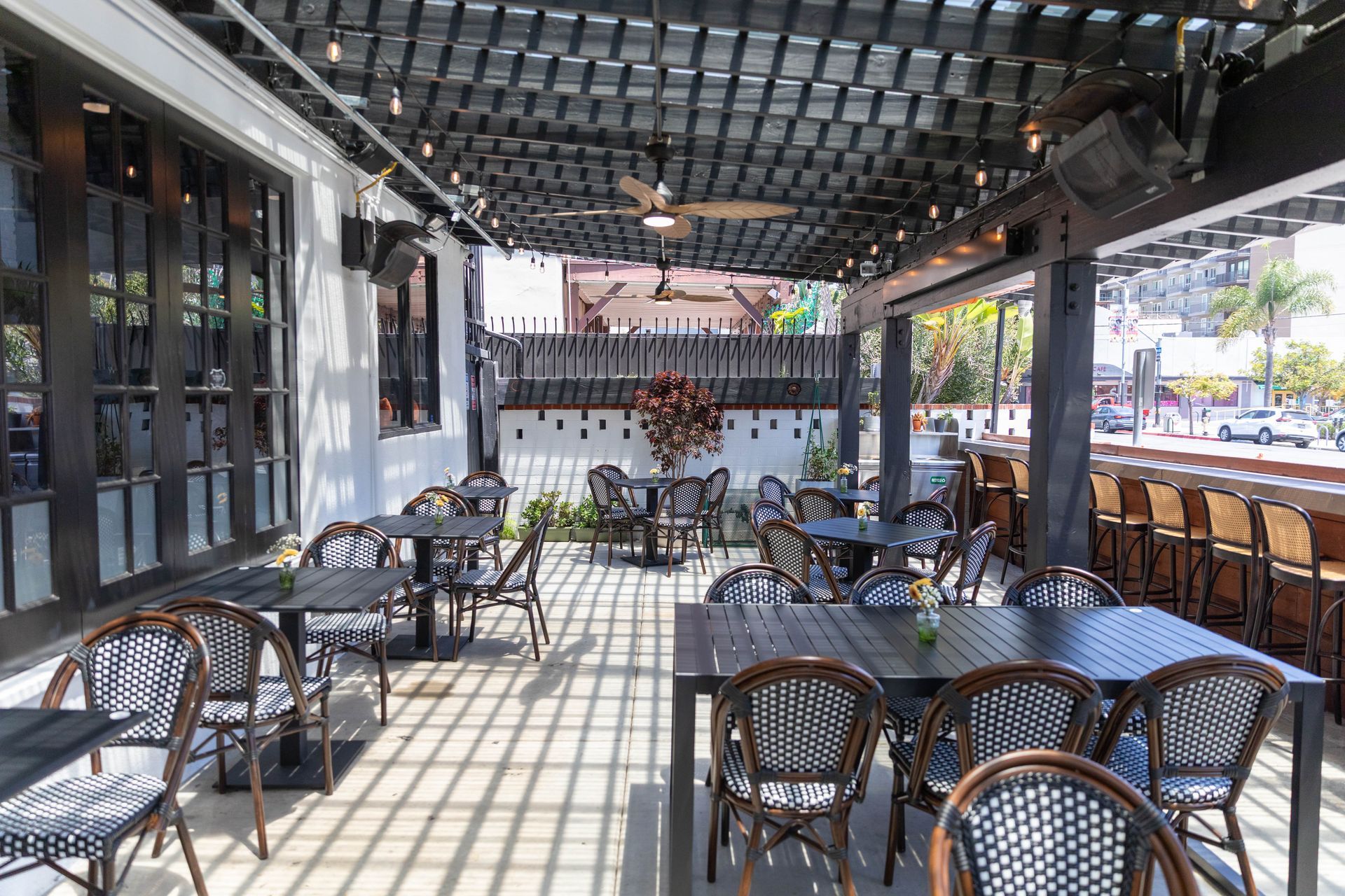 Outdoor café patio with black wicker tables and chairs under a shaded pergola, sunlight casting grid shadows.