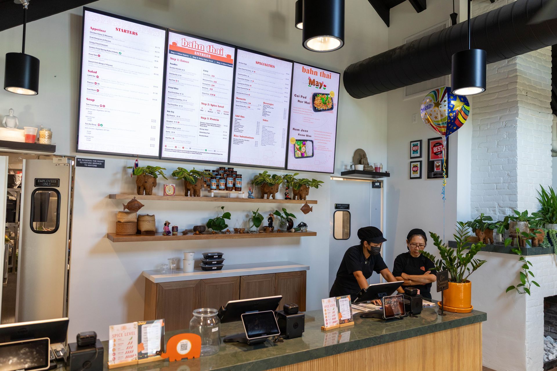 Cafe counter with two baristas, menu boards, hanging lights, and plants on shelves
