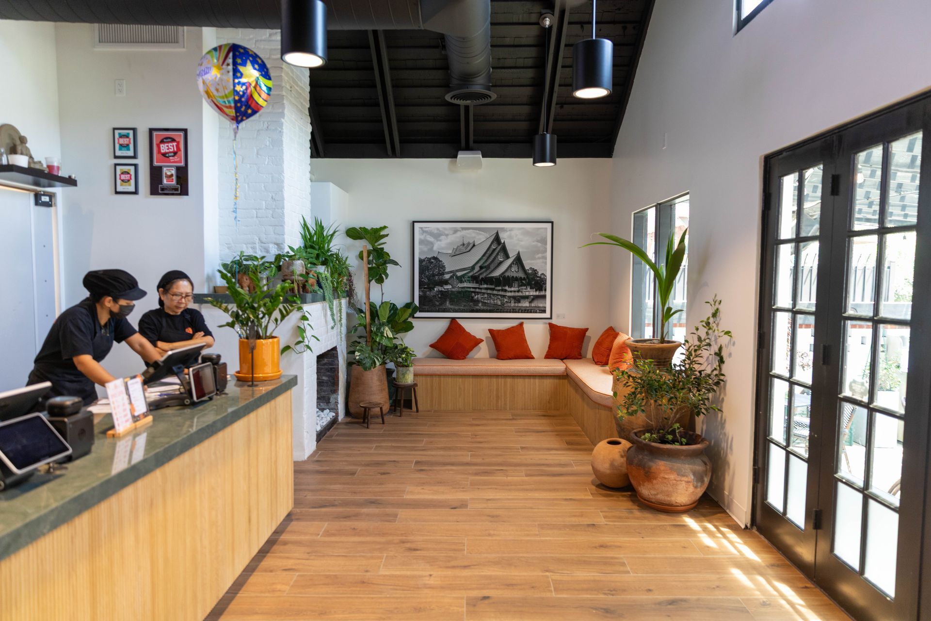Café interior with baristas at the counter, wooden seating, plants, and sunlight through glass doors