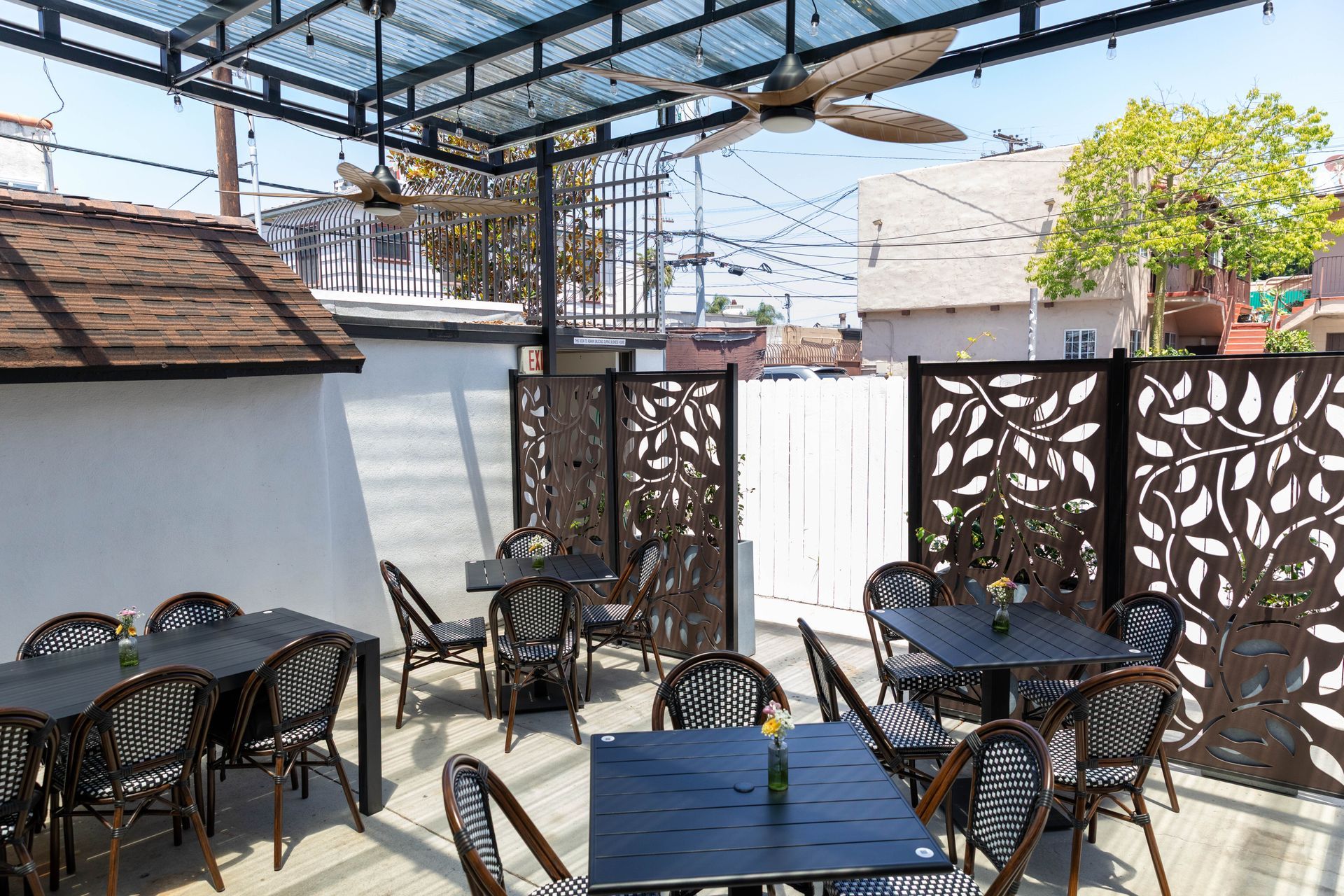 Outdoor café patio with black tables and chairs under a canopy, beside decorative privacy screens and a white wall