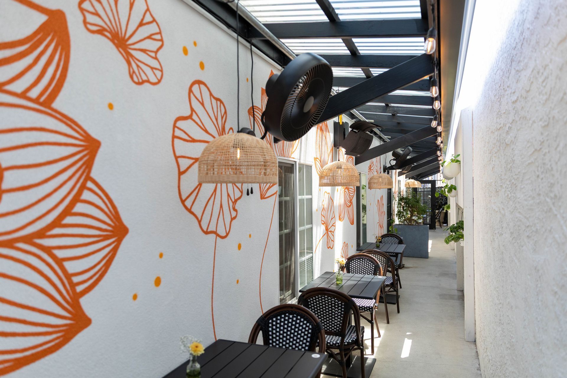 Outdoor café walkway with black chairs and tables beside a white wall with orange floral mural