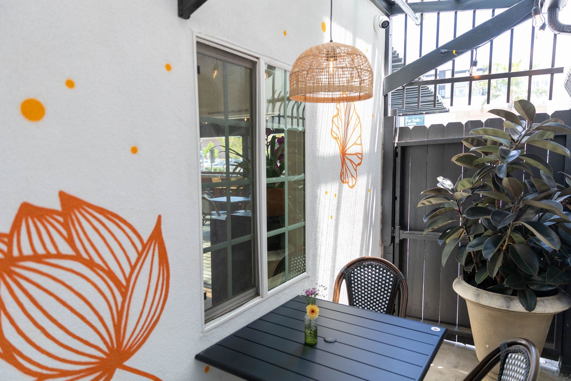 Patio seating area with a black table, woven lamp, potted plant, and orange leaf mural on a white wall