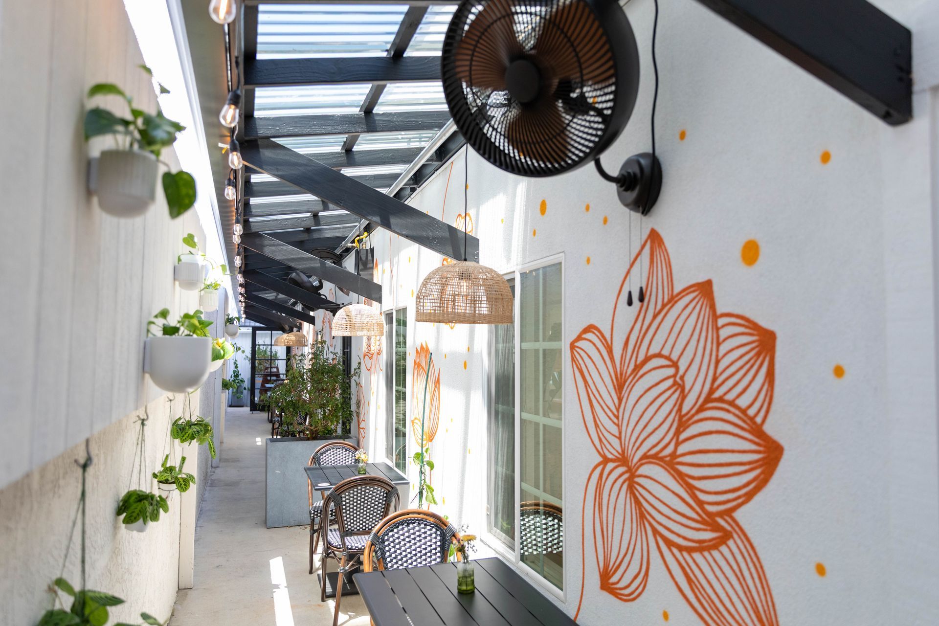Outdoor café corridor with potted plants, metal chairs, and orange floral wall art under fans