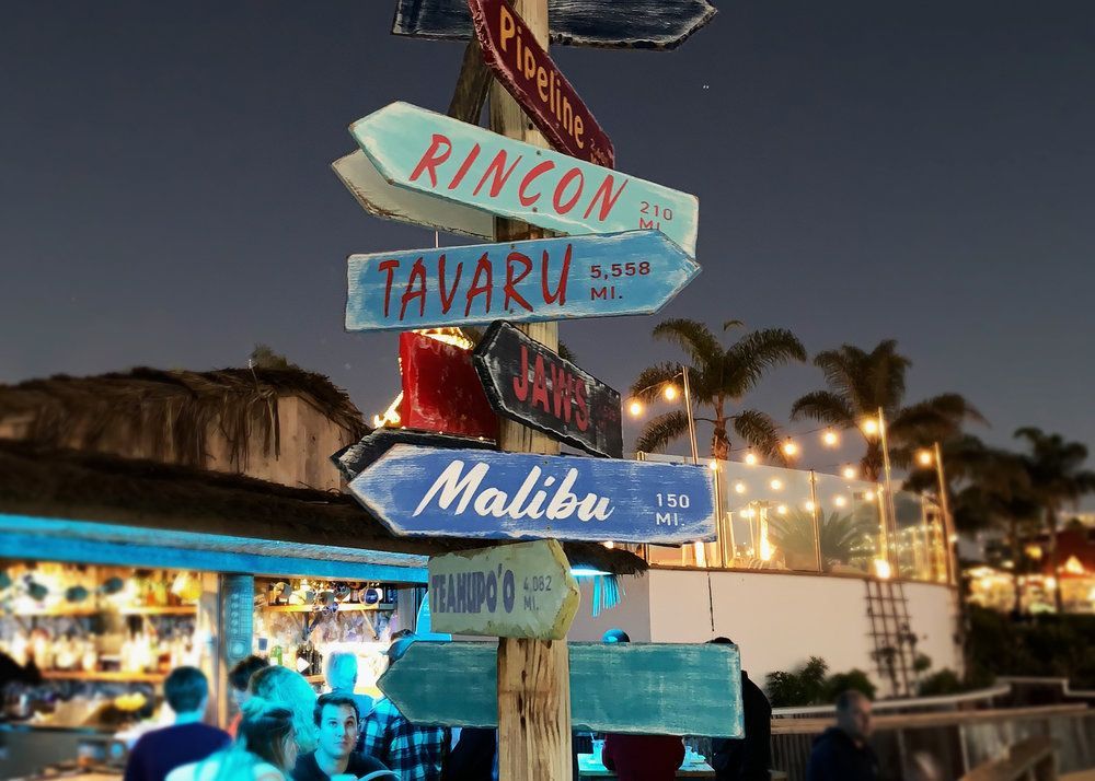 Colorful wooden signpost at a beach bar, with palm trees and string lights at dusk.