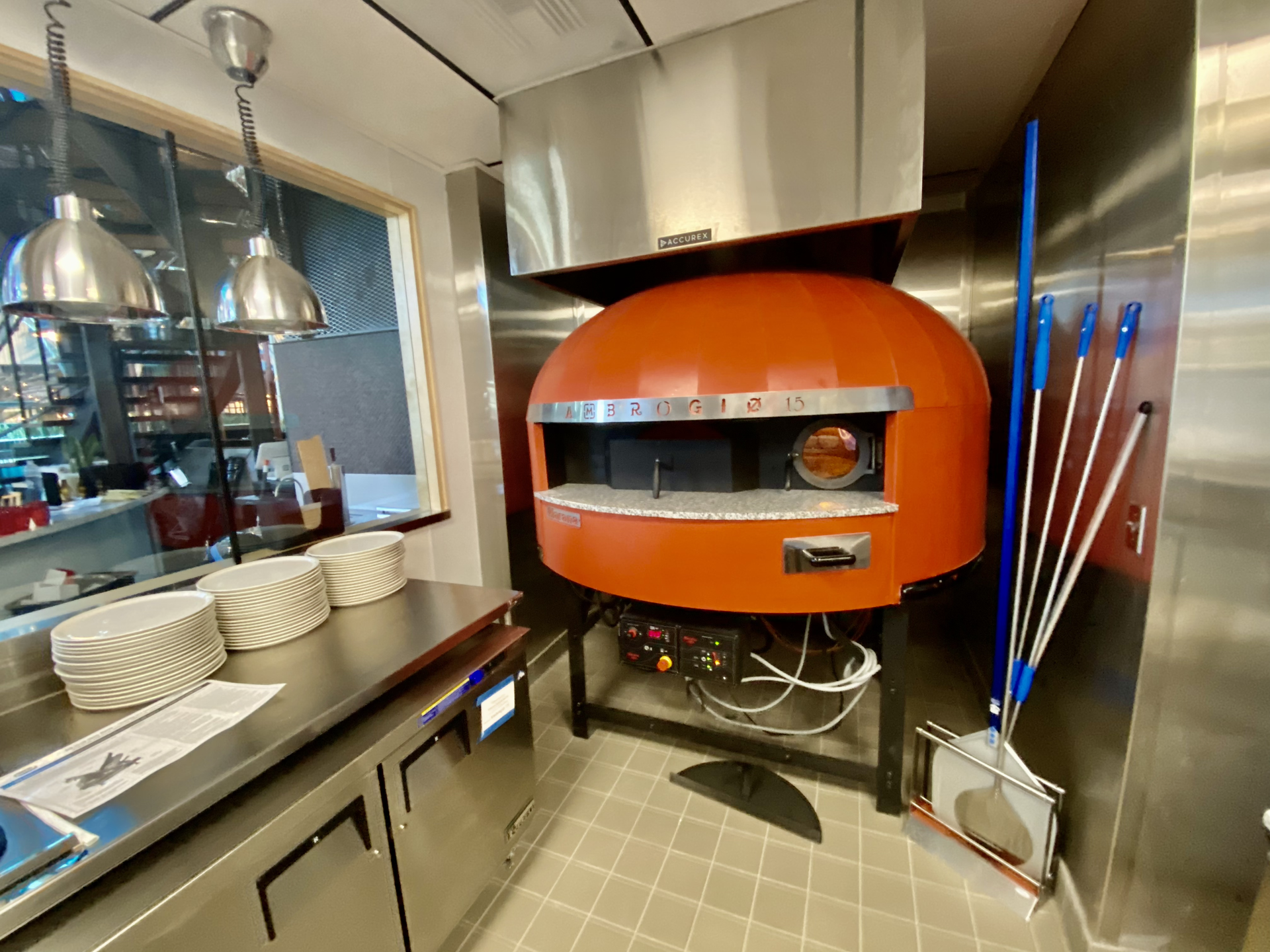 Orange pizza oven in a stainless-steel kitchen with stacked plates and cleaning tools nearby