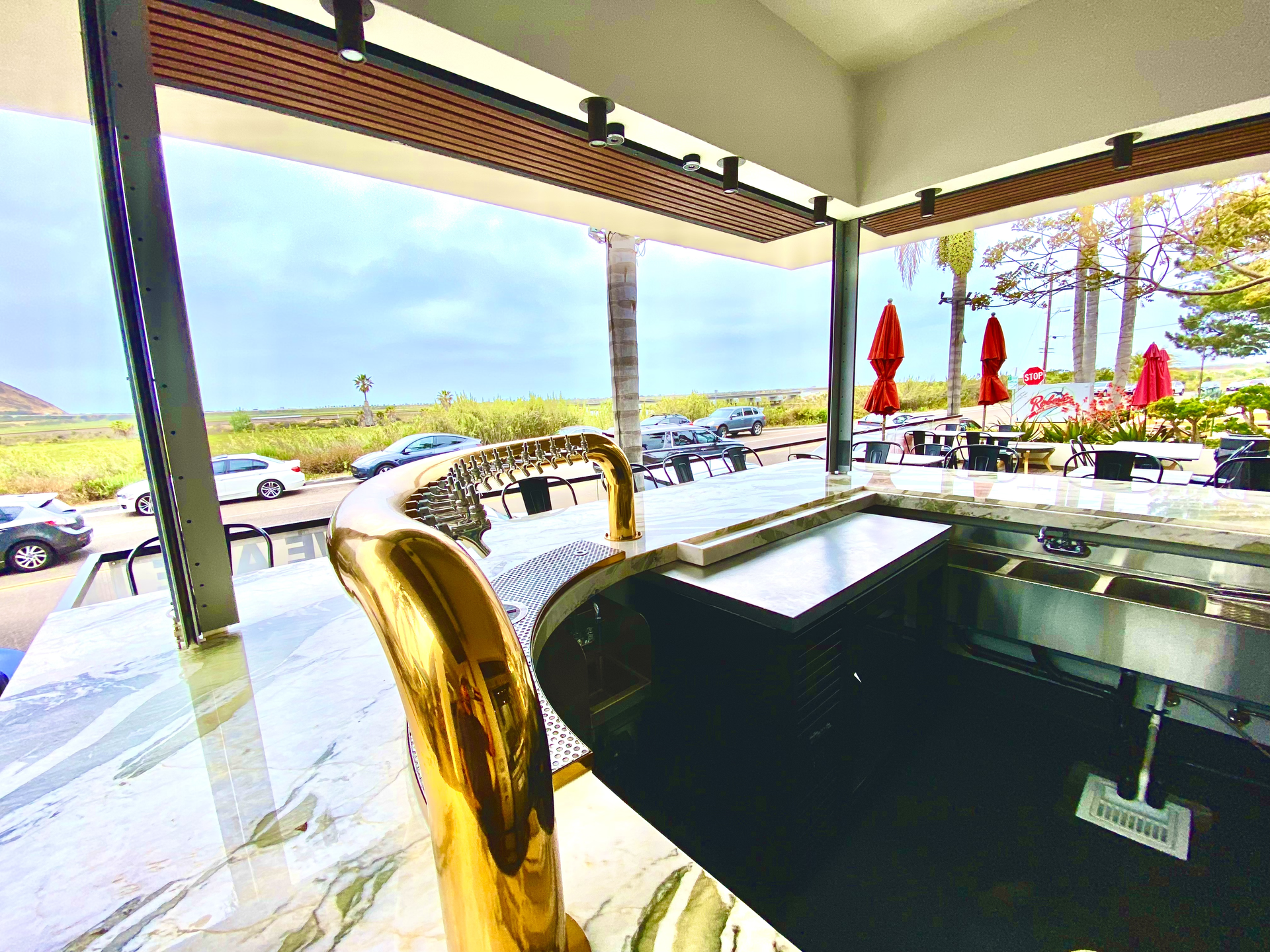 Outdoor hot tub under a shaded patio overlooking a pool and ocean view, with red umbrellas nearby