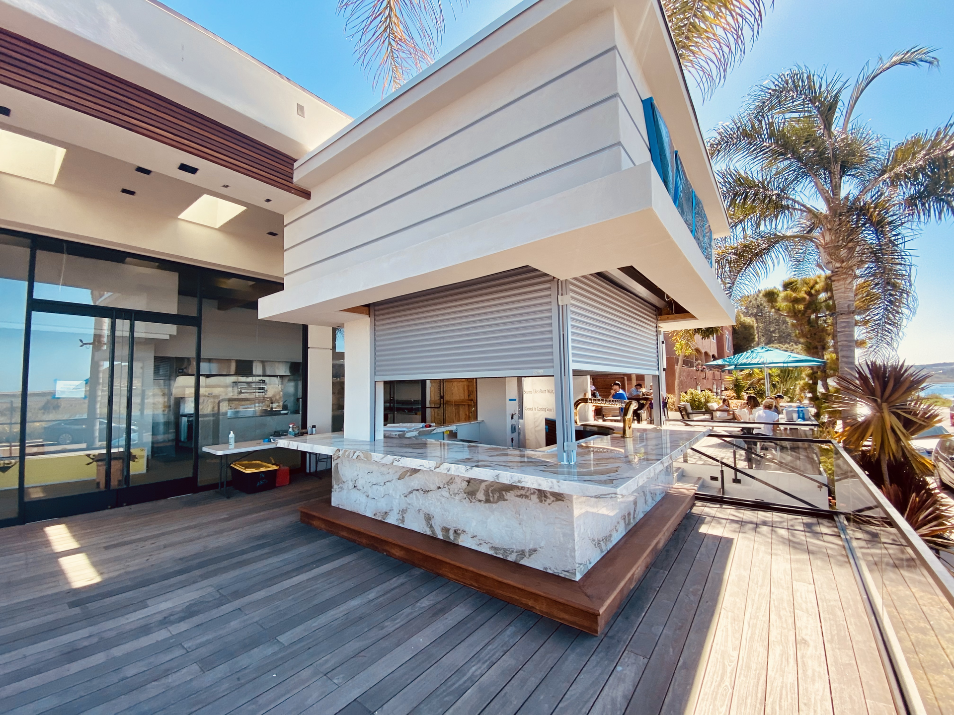 Modern rooftop bar with white canopy, wooden deck, glass walls, and palm trees in bright sunlight