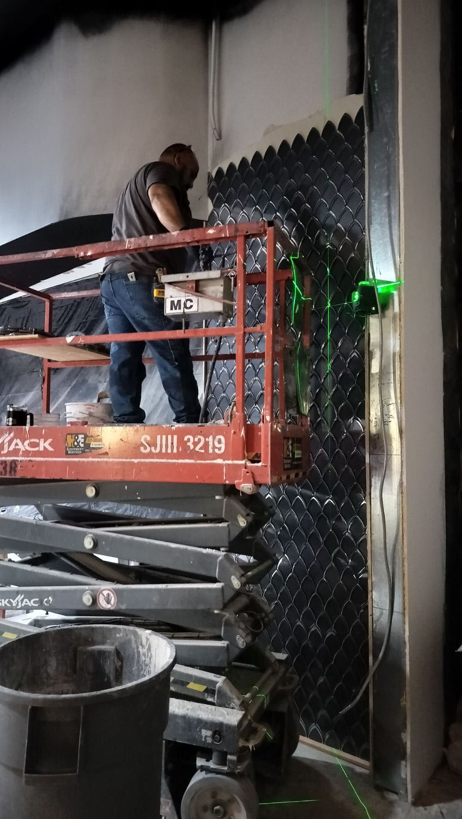 Worker on a red lift installing black wall panels in a narrow indoor space.